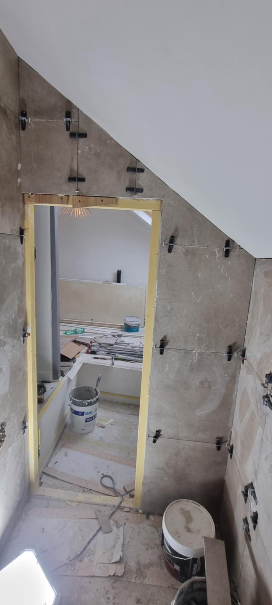 Ściana w trakcie wykańczania płytkami, z użyciem klipsów poziomujących. Widoczne wiaderka i narzędzia w tle, sufit ze skosem.