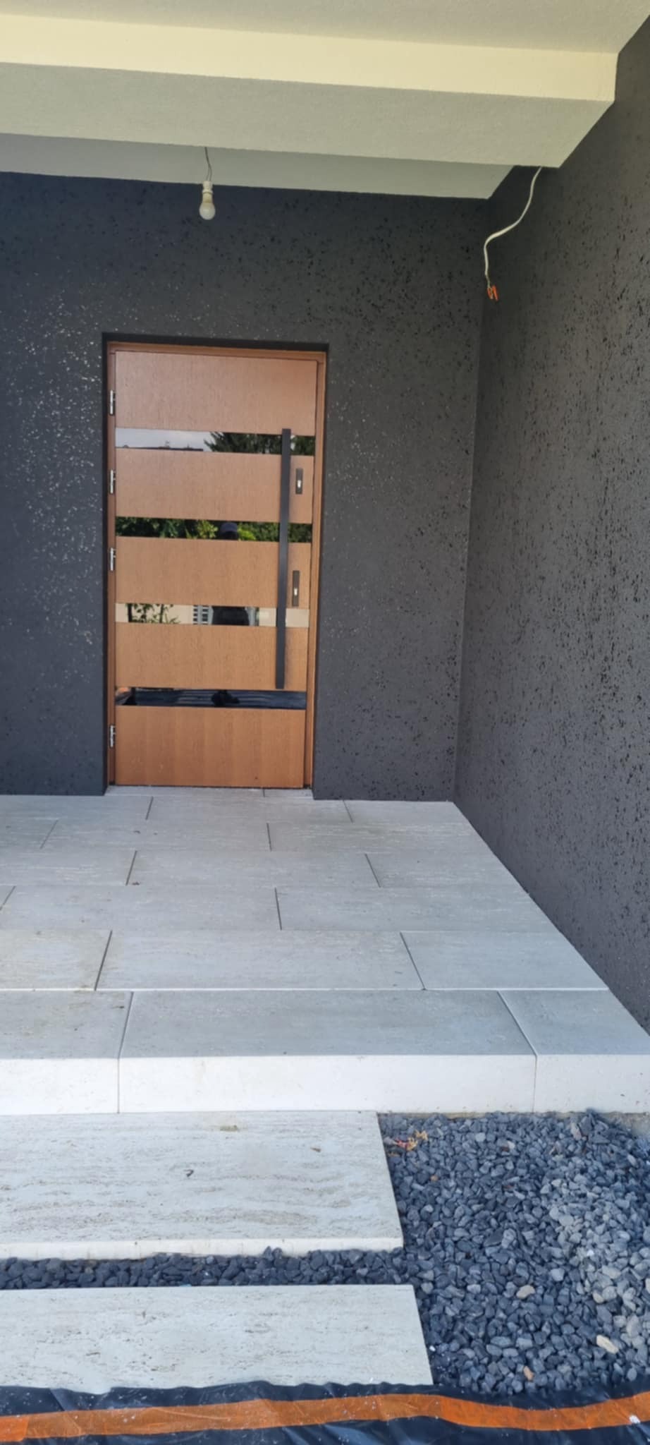 Nowoczesne drzwi wejściowe z poziomymi przeszkleniami, zamontowane w elewacji o szarej, ziarnistej fakturze. Jasne płytki na podeście i drobny żwir u podstawy.