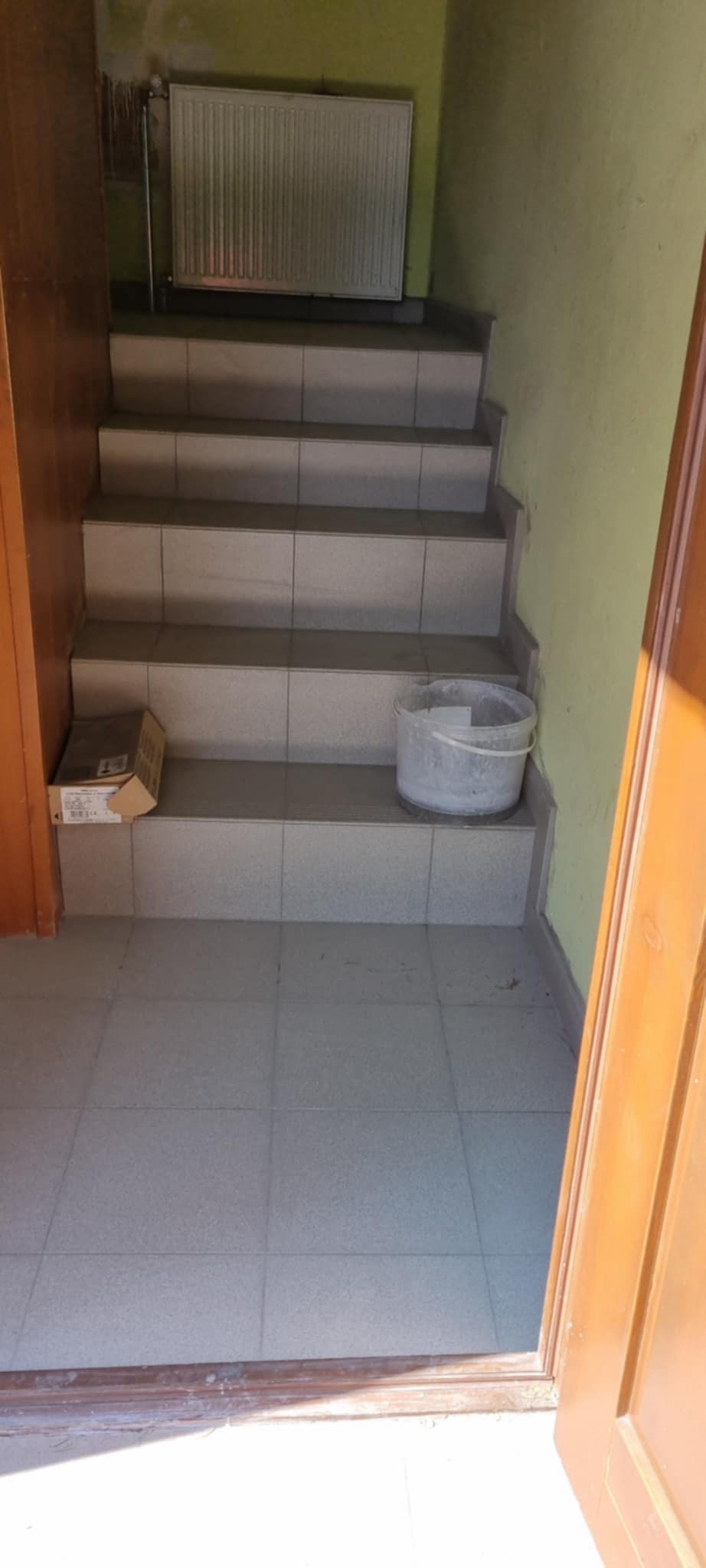 Wyłożone płytkami schody wewnątrz budynku, widoczny grzejnik na ścianie, karton i wiadro na schodach. Ujęcie z dołu schodów, widoczne otwarte drzwi.