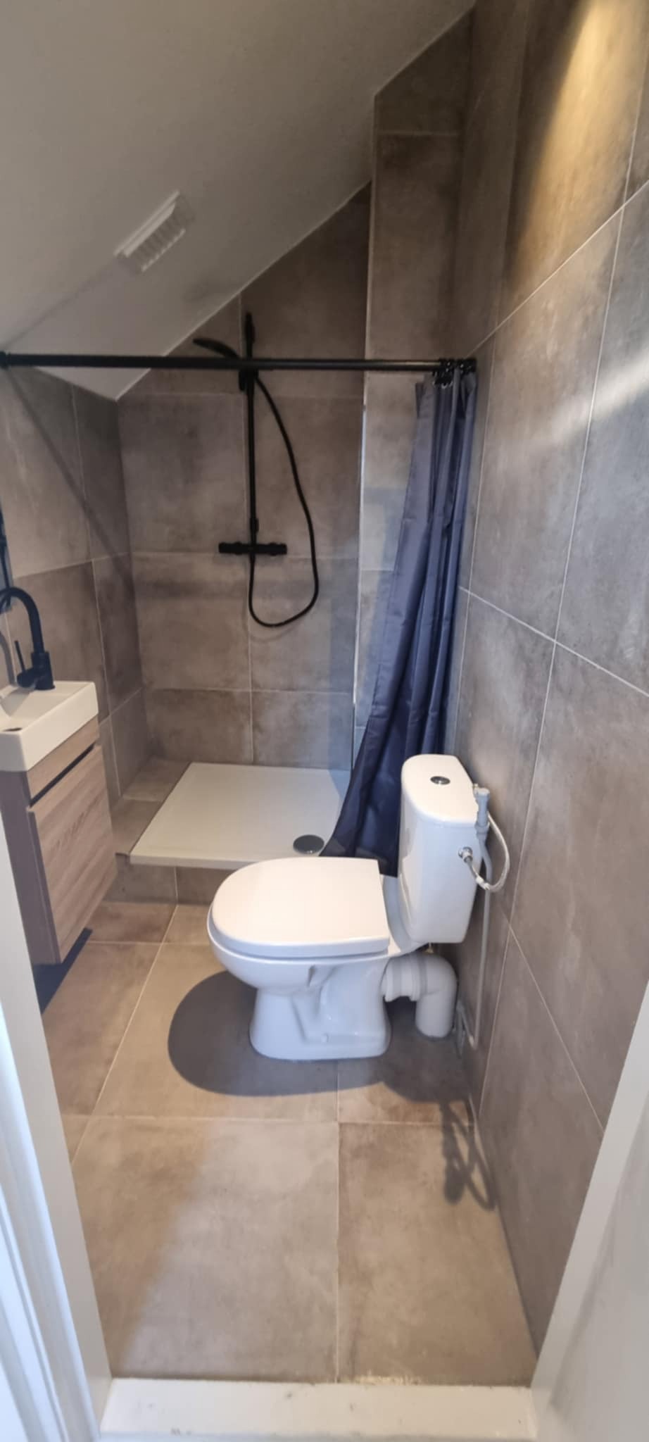 Nowoczesna łazienka w szarościach: kabina prysznicowa typu walk-in, podwieszana toaleta z bidetką i umywalka z szafką. Uwagę zwraca czarna armatura i oświetlenie.