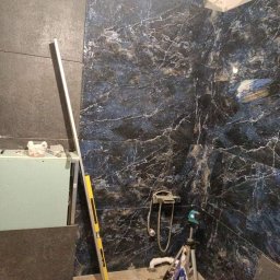 Rafał Szelągowski - Ściana w łazience wyłożona ciemnymi płytkami imitującymi marmur z białymi żyłami. Widoczne narzędzia glazurnicze: poziomica, laser krzyżowy, paczka kleju.