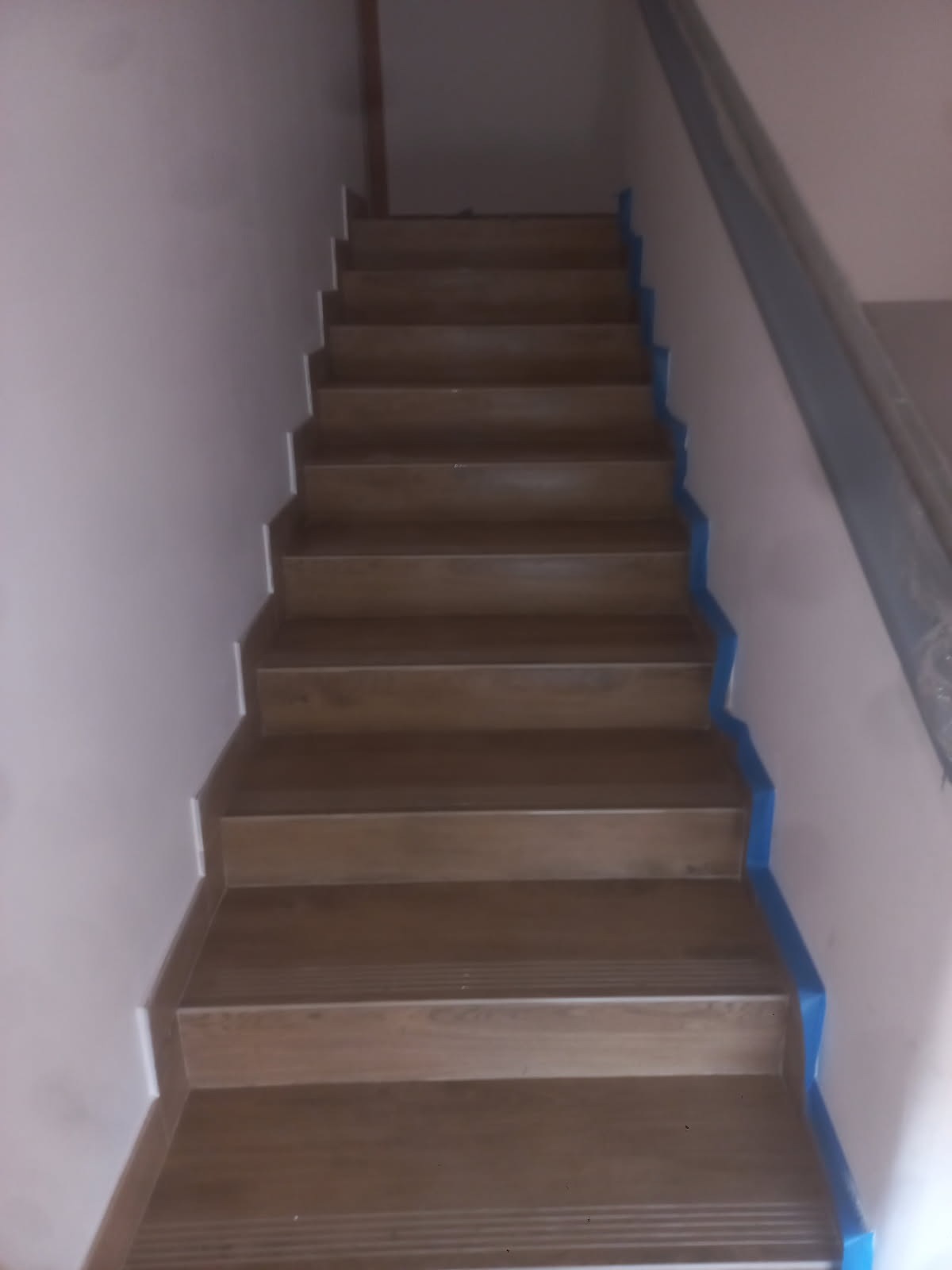 Wykończone schody z brązowych płytek, zabezpieczone niebieską taśmą malarską przy ścianie. Widok z dołu, schody prowadzą do góry.
