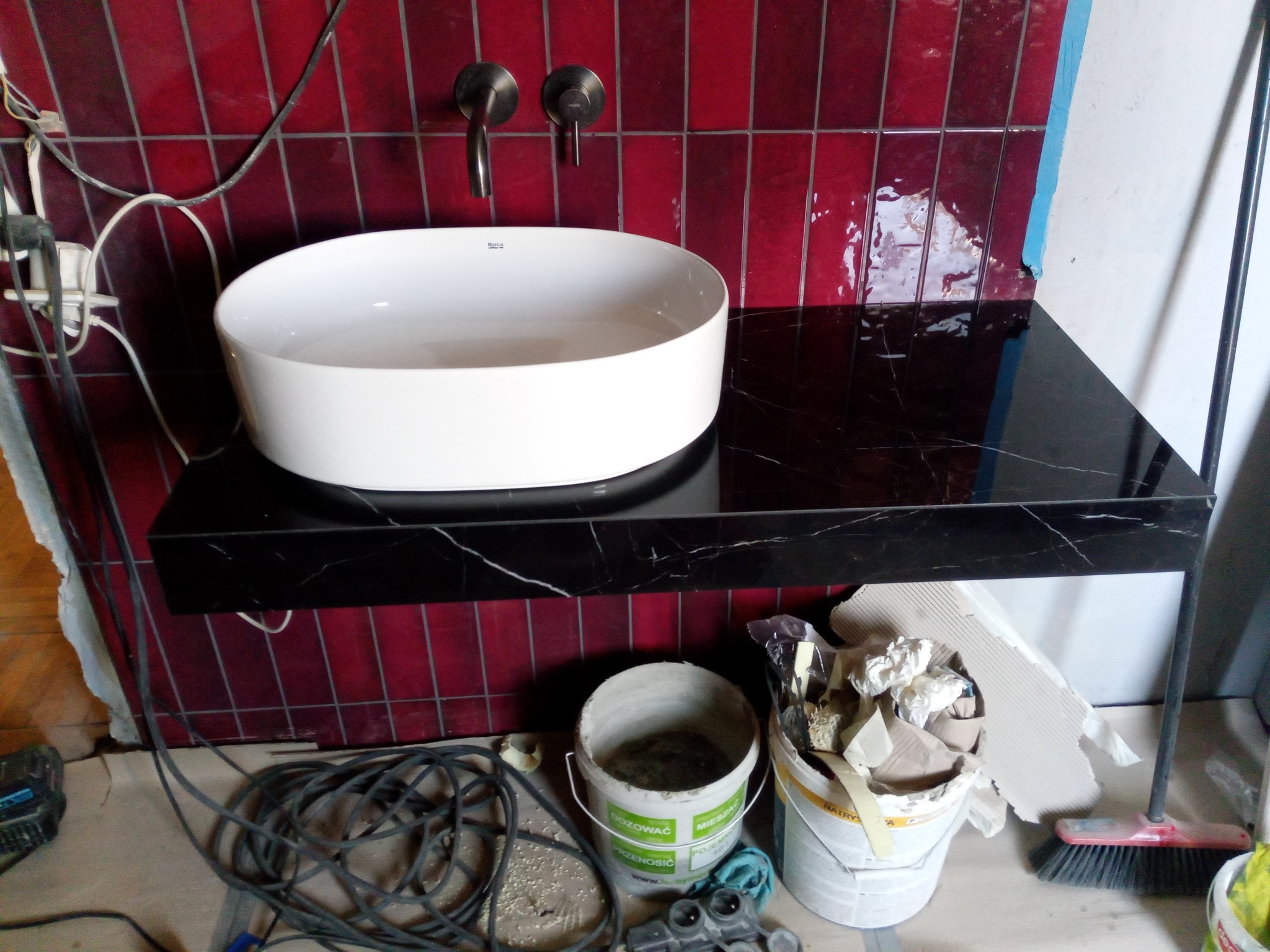 Łazienka w trakcie remontu: biała umywalka Roca na czarnym blacie z marmuru, czerwone płytki na ścianie, widoczne narzędzia i materiały budowlane.