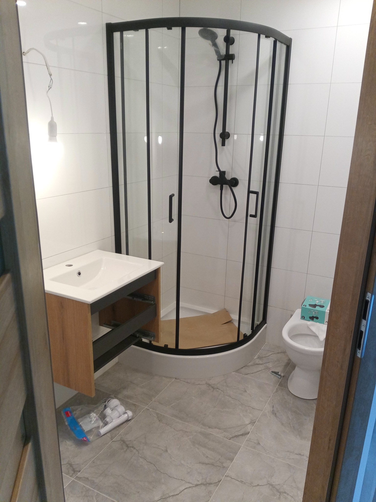 Nowoczesna łazienka po remoncie: kabina prysznicowa z czarną armaturą, umywalka z szafką w odcieniach drewna, biała toaleta i szare płytki podłogowe.