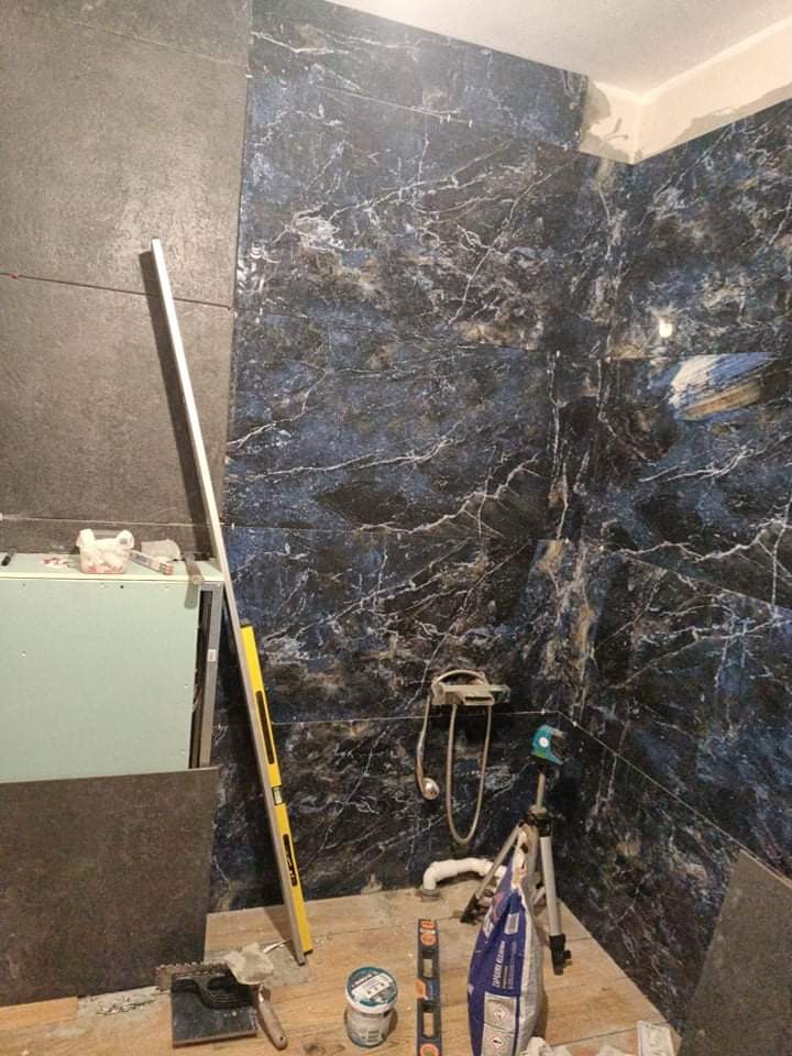 Ściana w łazience wyłożona ciemnymi płytkami imitującymi marmur z białymi żyłami. Widoczne narzędzia glazurnicze: poziomica, laser krzyżowy, paczka kleju.