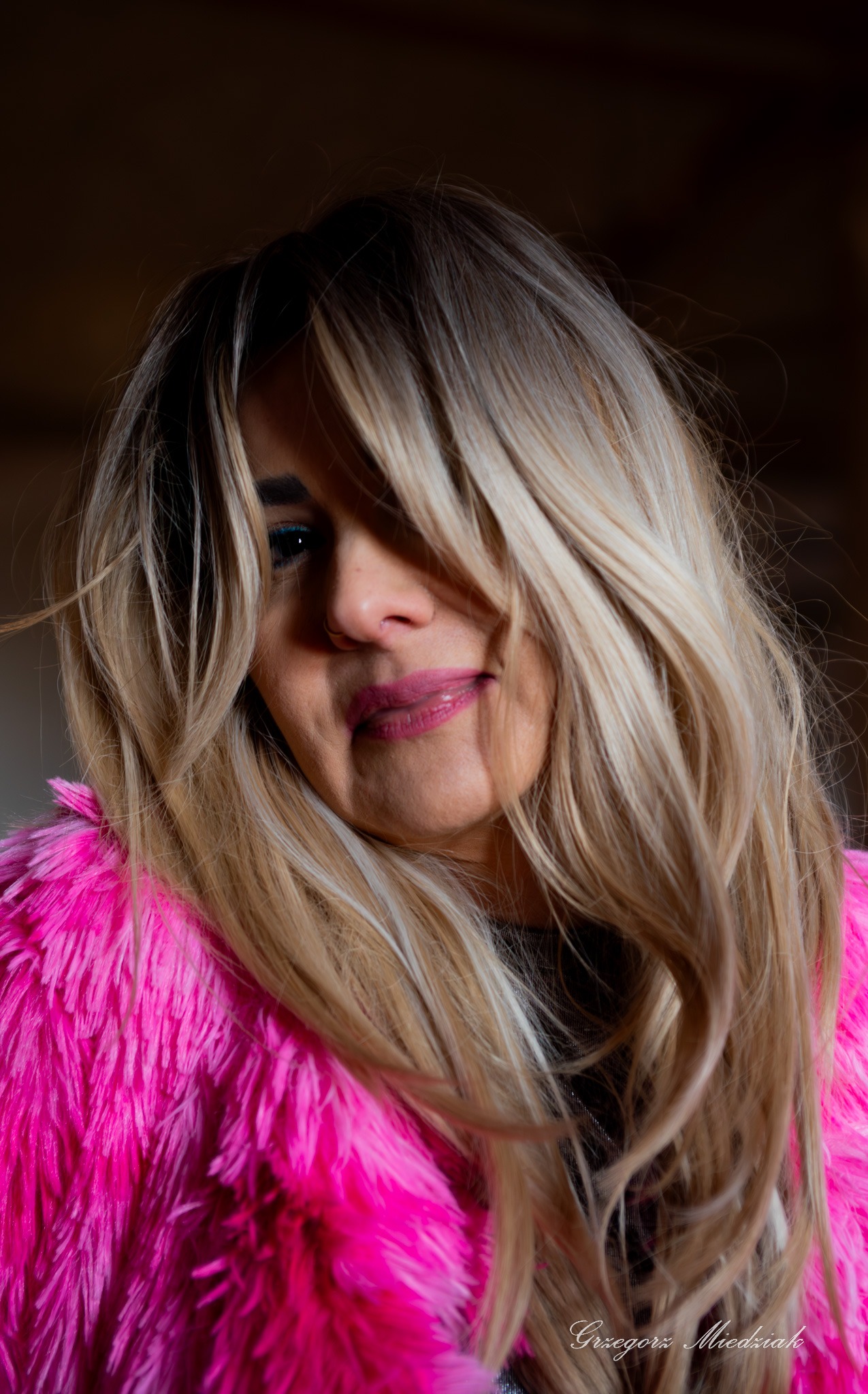 Portret kobiety z blond włosami częściowo zasłaniającymi twarz, ubrana w różowe futro. Widoczny makijaż i delikatny uśmiech. Zdjęcie z sygnaturą fotografa.
