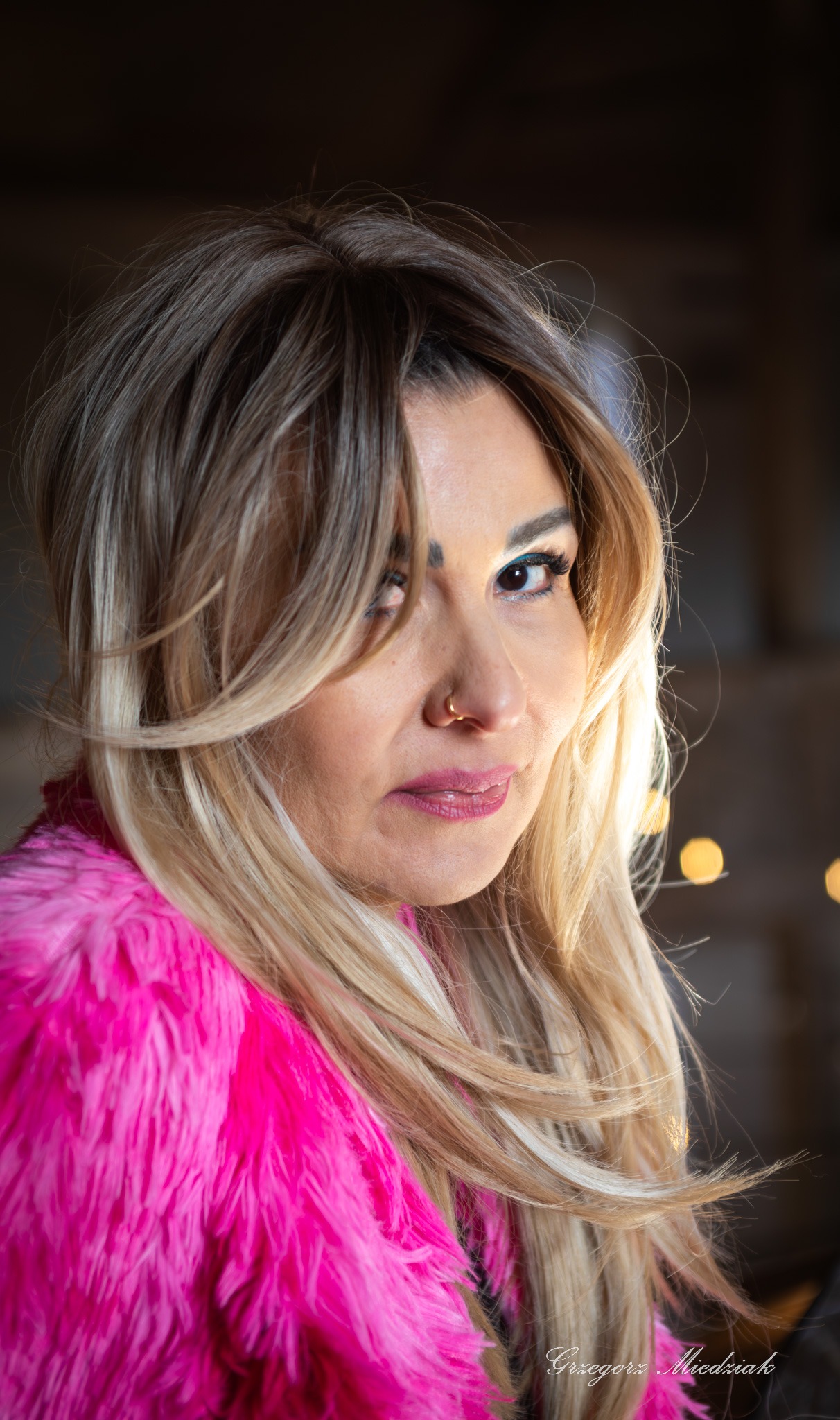 Portret kobiety z blond włosami i różowym futrem, z kolczykiem w nosie, spoglądającej w bok na rozmytym tle z lampkami.