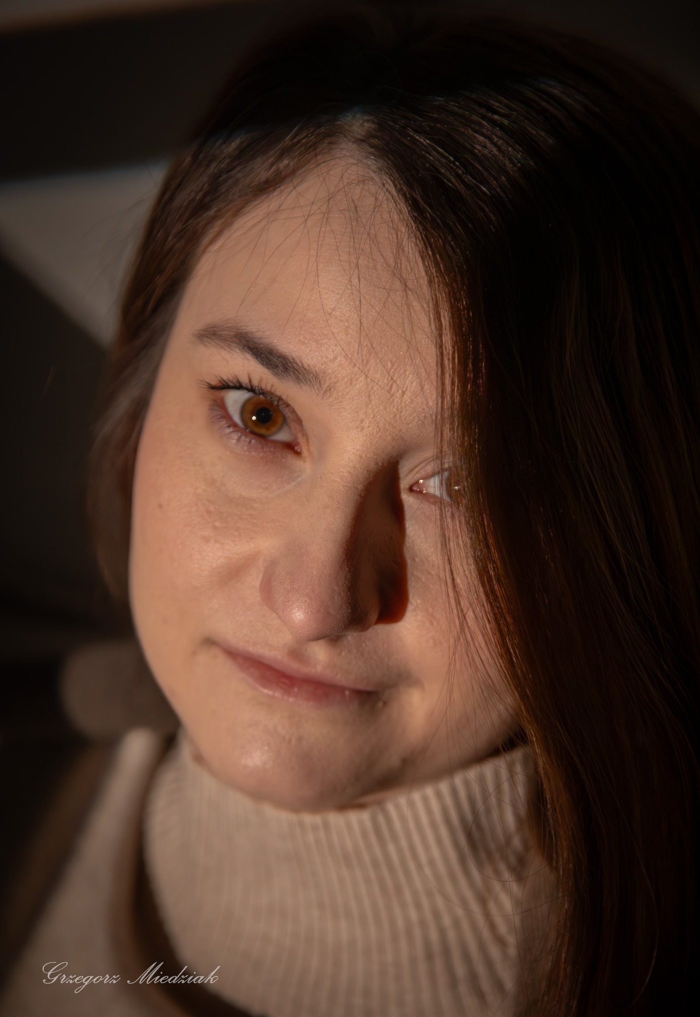 Portret kobiety z brązowymi włosami i bursztynowymi oczami, oświetlonej bocznym światłem, w beżowym golfie. Ujęcie z dołu, podkreślające rysy twarzy.