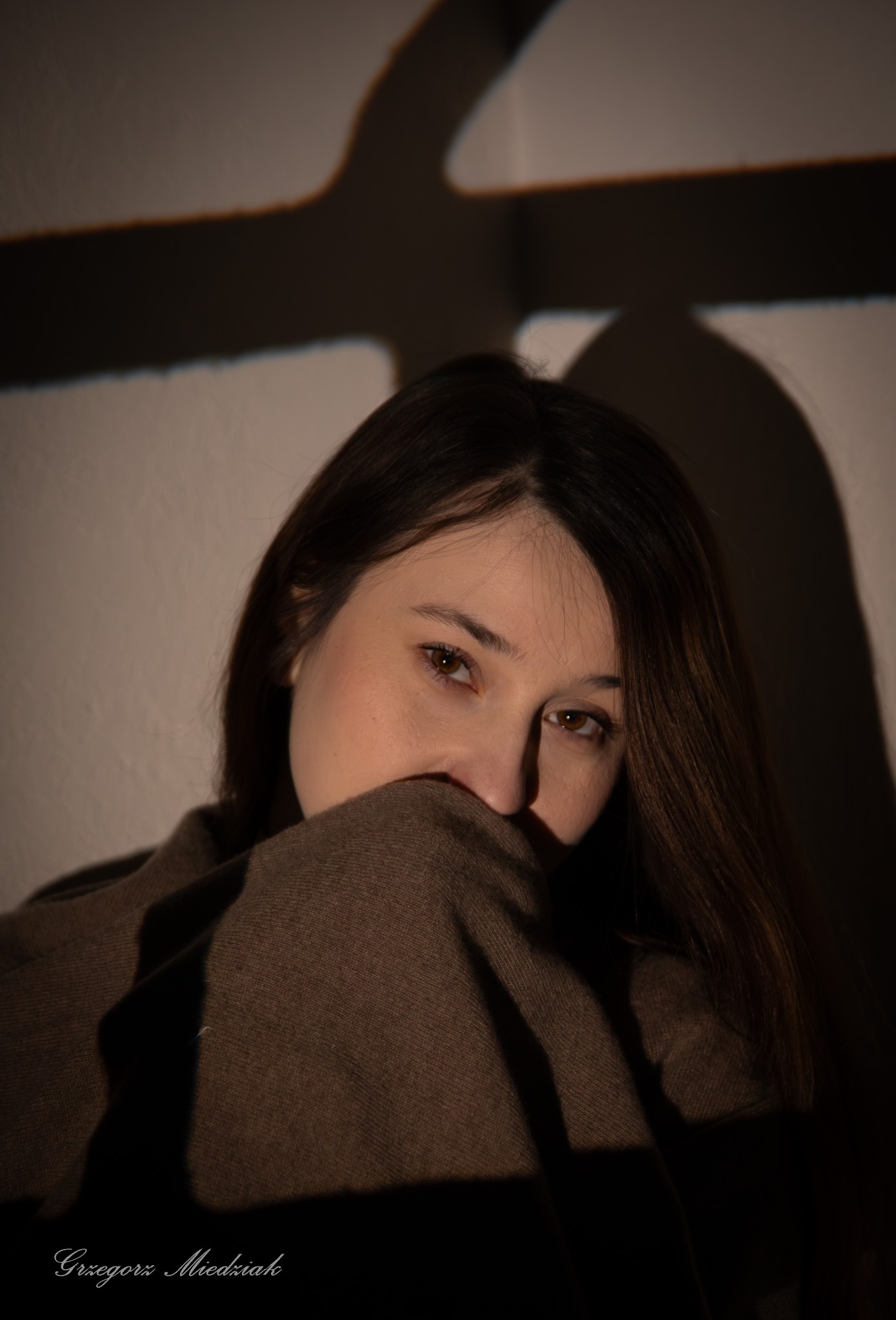 Portret kobiety z brązowymi włosami, częściowo zasłoniętej materiałem w ciepłym oświetleniu z cieniem krzyża na ścianie. Ujęcie z bliska, skupione na twarzy.