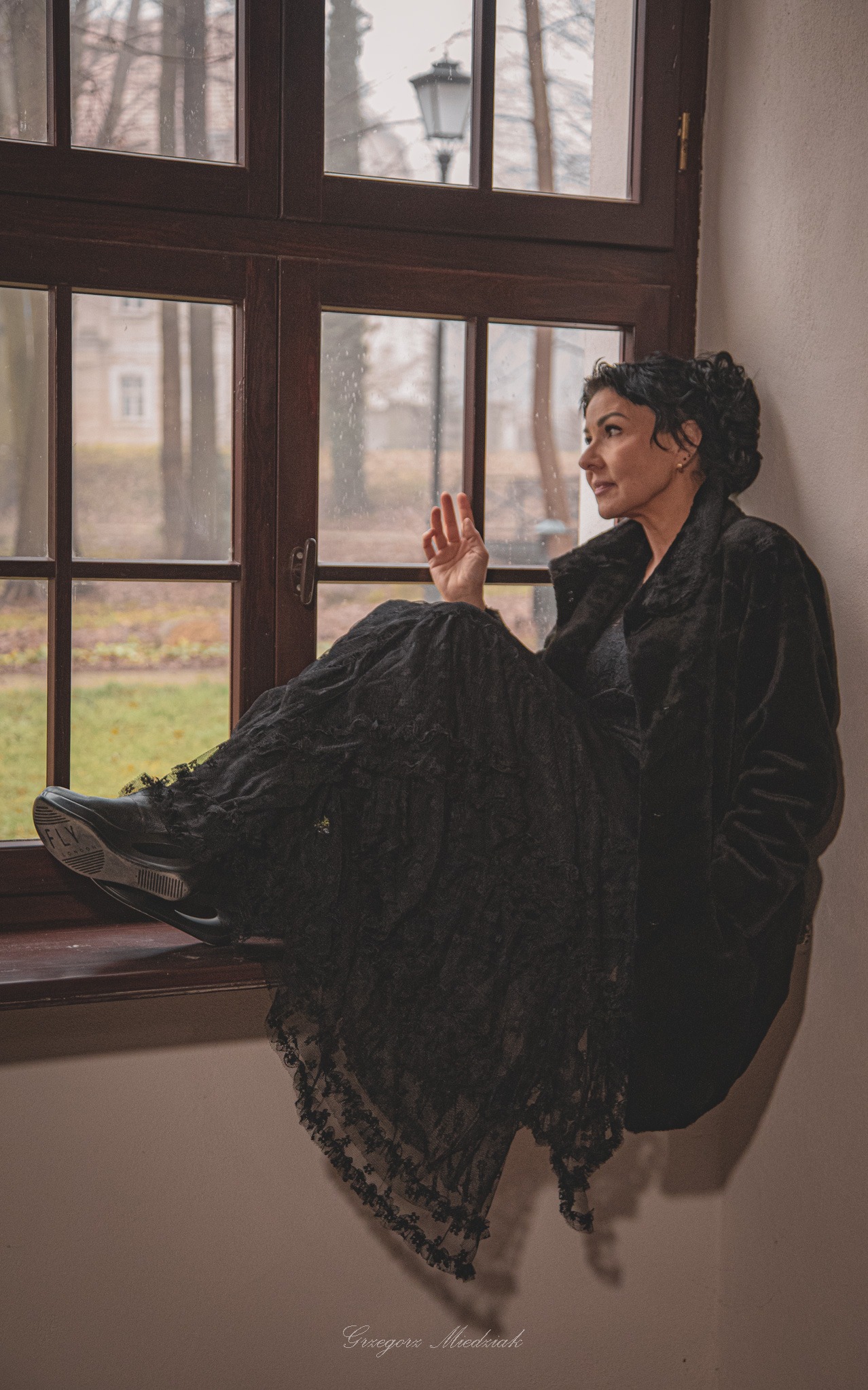 Kobieta w czarnej sukni i płaszczu siedzi na parapecie okna, spoglądając na zewnątrz. Widoczny budynek i latarnia za oknem. Stylizowane ujęcie.