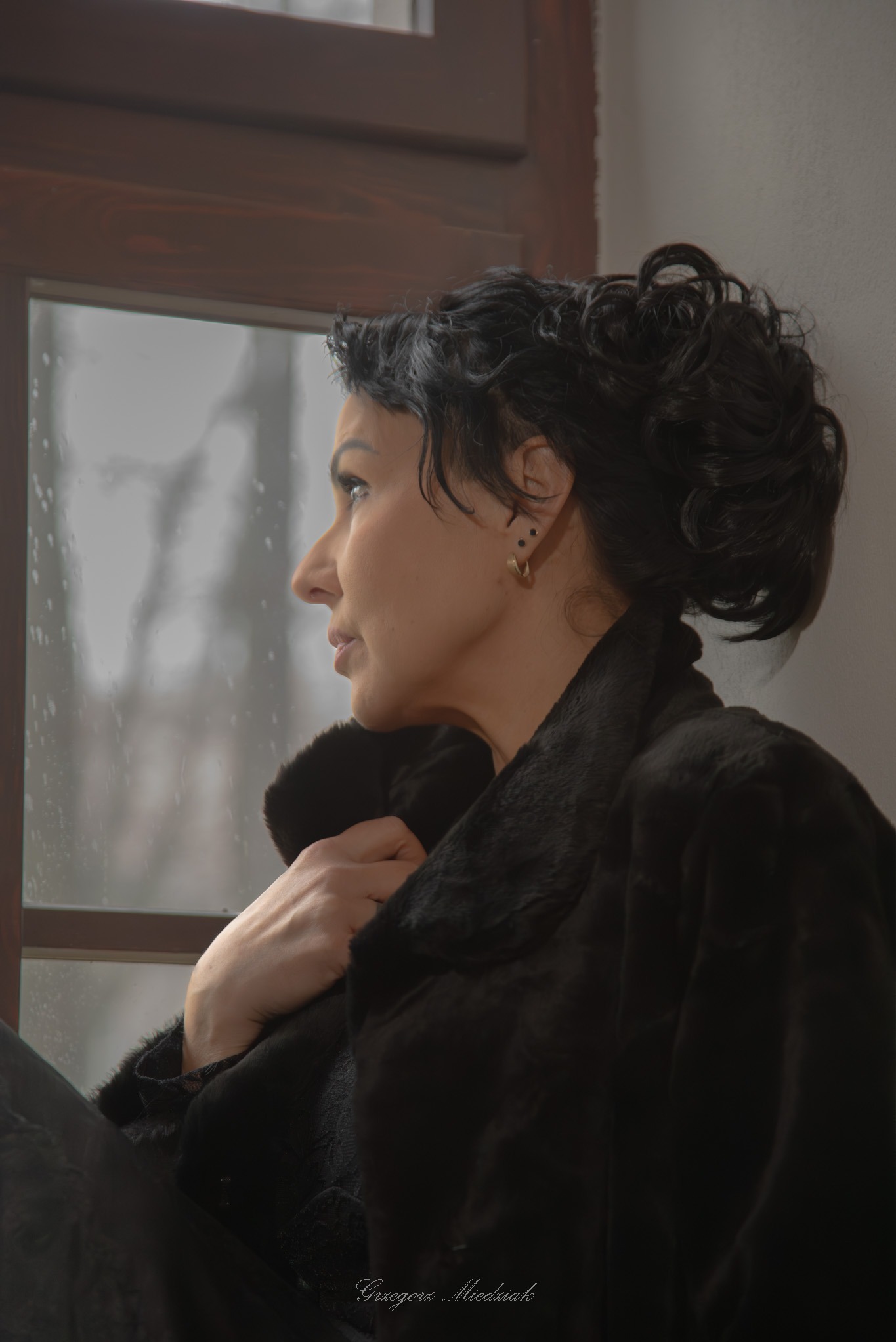 Portret kobiety w futrze, spoglądającej przez okno z kroplami deszczu. Profil, stonowana kolorystyka, miękkie światło. Widoczne kolczyki i upięte włosy.