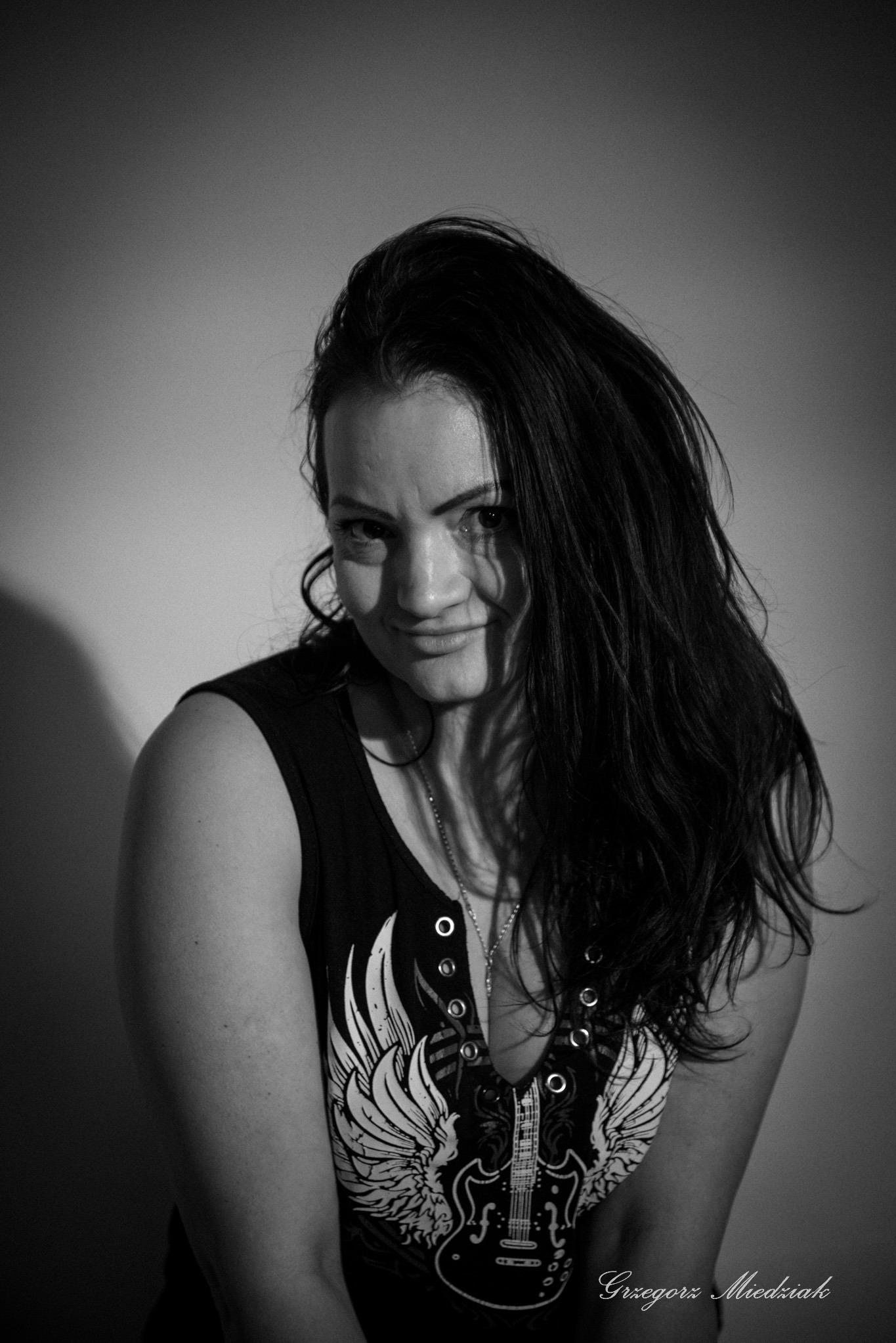 Czarno-biały portret kobiety z długimi włosami, ubranej w koszulkę z motywem gitary i skrzydłami, oświetlonej z boku, tworząc cień na ścianie. Wyraz twarzy kokieteryjny.