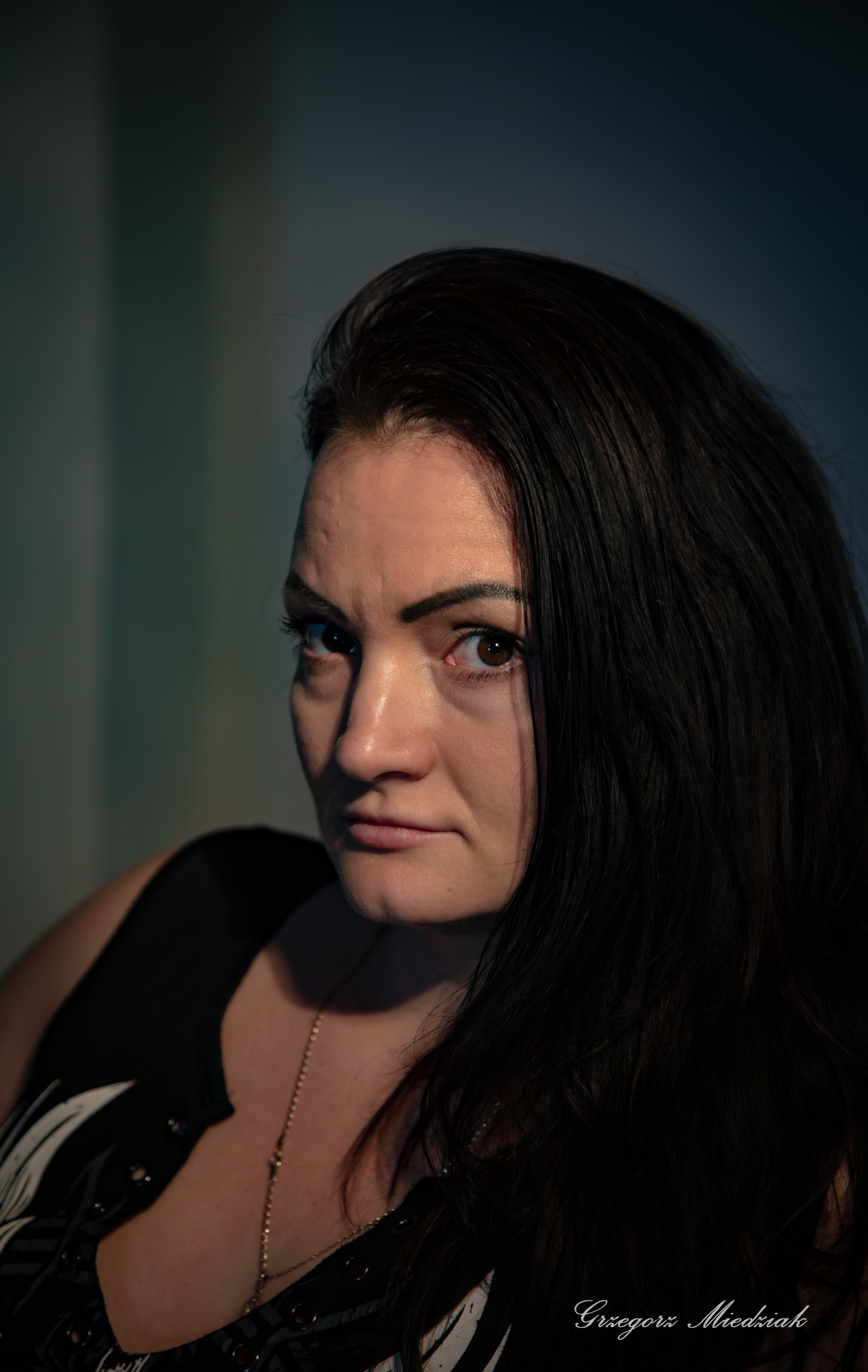 Portret kobiety o ciemnych włosach, z delikatnym makijażem i złotym łańcuszkiem, uchwycony w półmroku z subtelnym oświetleniem akcentującym rysy twarzy.