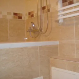 Beżowe płytki w łazience z dekoracyjnym pasem, bateria wannowa z prysznicem i grzejnik drabinkowy.