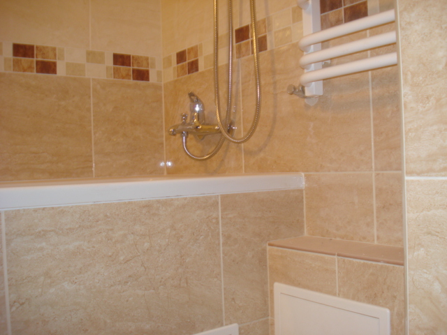 Beżowe płytki w łazience z dekoracyjnym pasem, bateria wannowa z prysznicem i grzejnik drabinkowy.