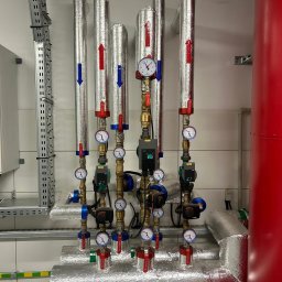 Hydraulika od A do Z - Izolowane rury instalacji grzewczej z manometrami i zaworami, widoczne strzałki kierunku przepływu, na tle białych płytek i czerwonego zbiornika.