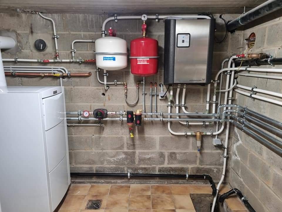 Złożona instalacja grzewcza w piwnicy: biały piec, srebrny panel sterowania, czerwony i biały zbiornik wyrównawczy, liczne rury i zawory na tle surowej ściany z cegły.