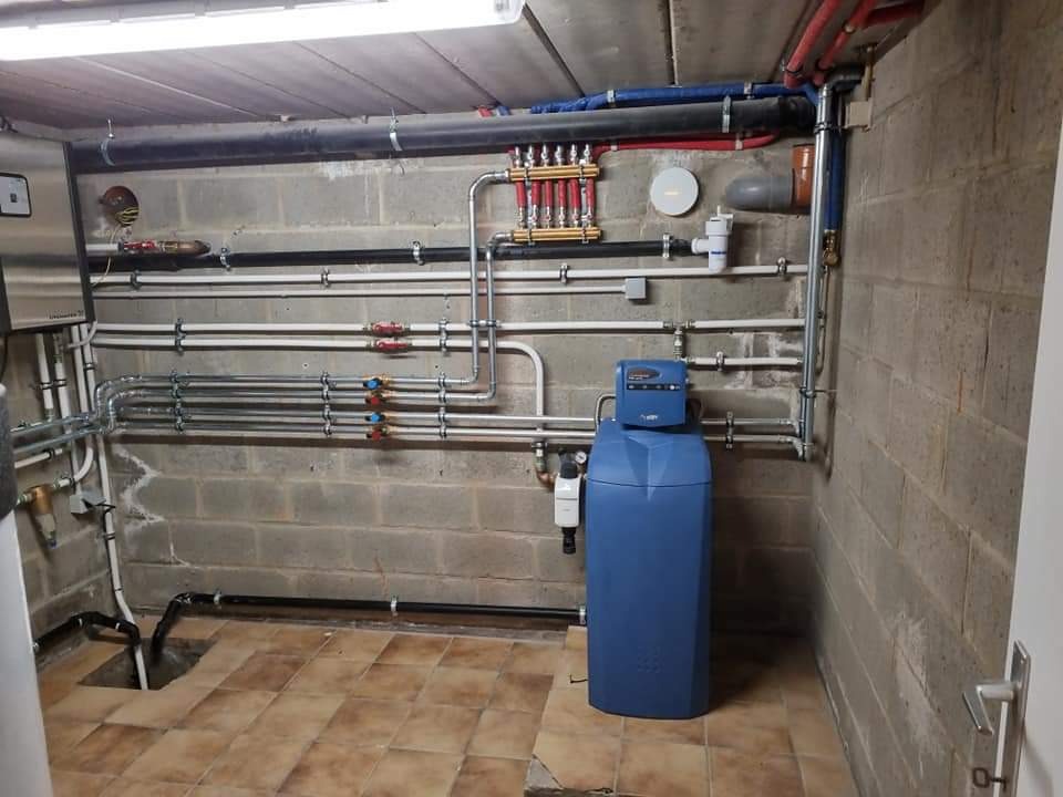 Piwnica z rozbudowaną instalacją hydrauliczną: rury, zawory, rozdzielacz, zmiękczacz wody. Widoczne elementy kotłowni i instalacji kanalizacyjnej.