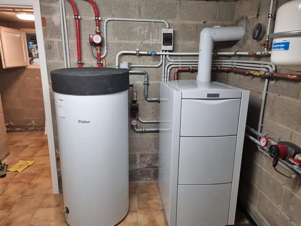 Biały piec CO Vaillant z zasobnikiem ciepłej wody w kotłowni z instalacją grzewczą. Widoczne rury miedziane i plastikowe, pompy obiegowe. Ściany z surowego betonu.