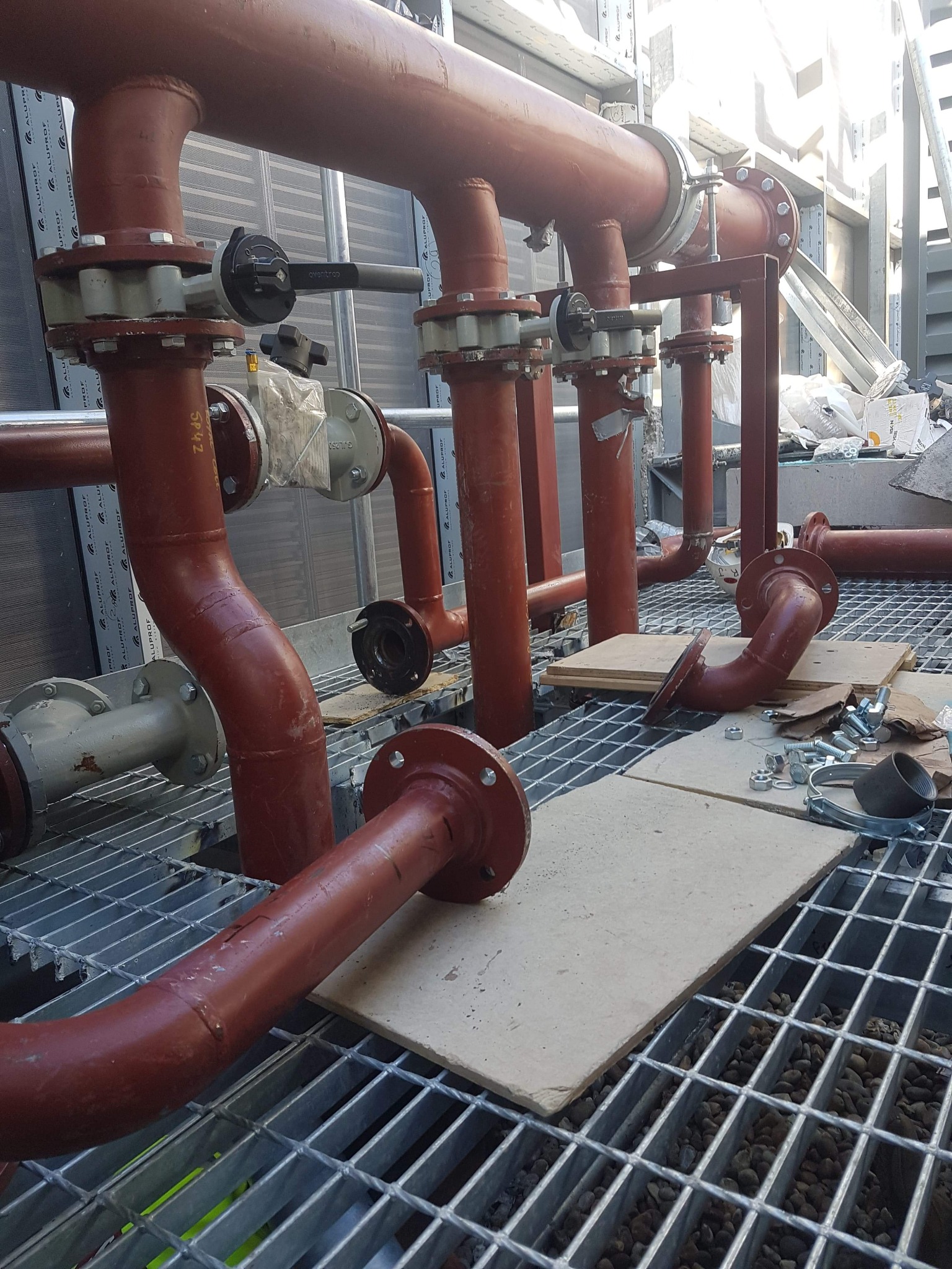 Czerwone rury instalacyjne z zaworami i elementami montażowymi na metalowej kratownicy. Widoczne łączenia kołnierzowe i fragmenty izolacji. Częściowo zdemontowana instalacja.