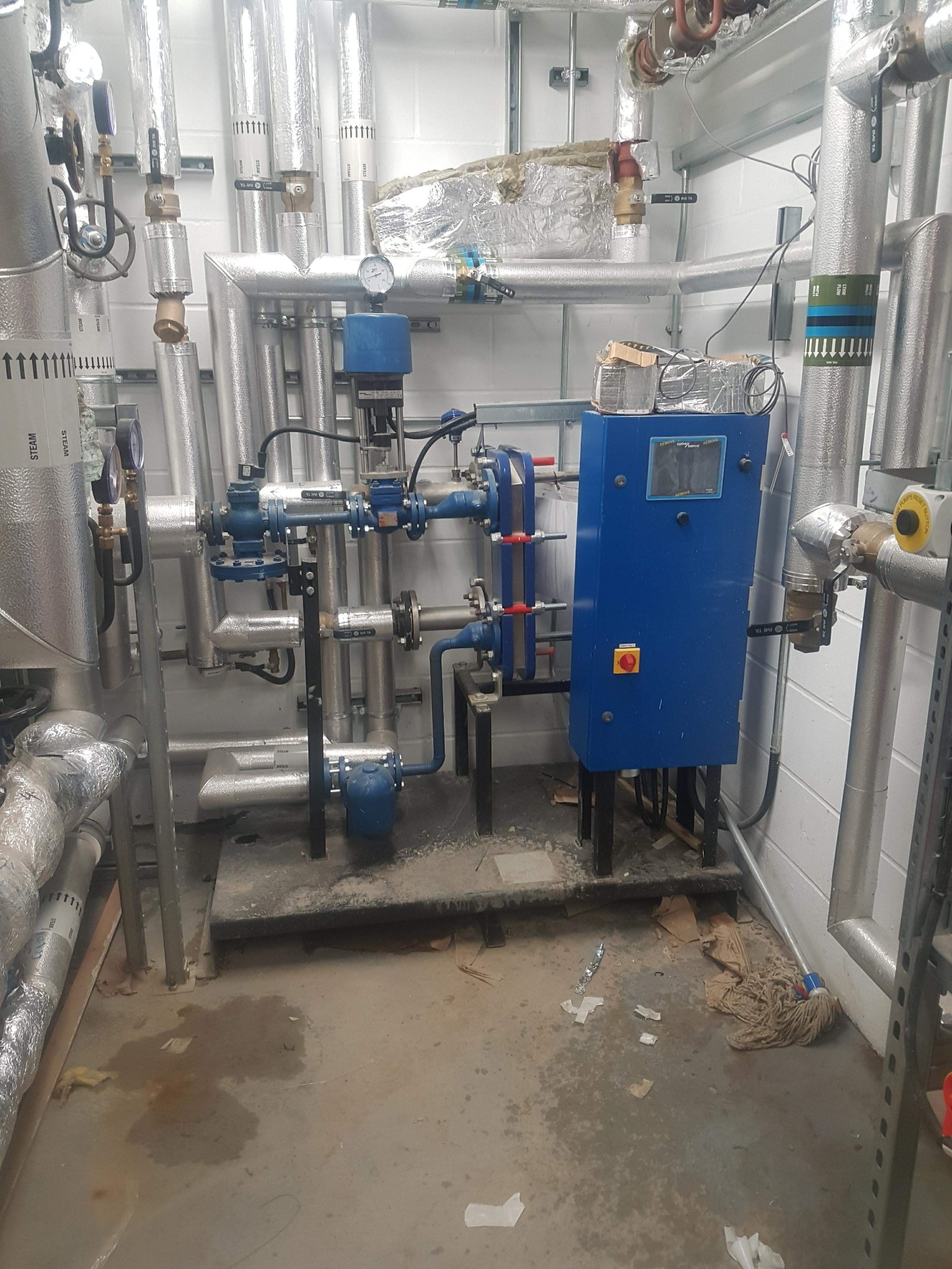 Węzeł cieplny z niebieską szafą sterowniczą, rurami w izolacji i manometrami. Widoczne zawory, pompy i instalacja. Podłoga zabrudzona.
