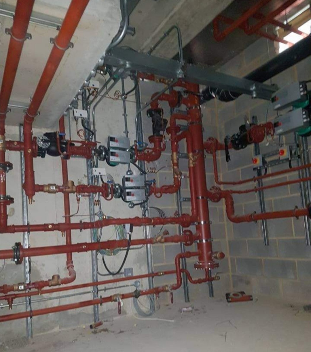 Rozbudowana instalacja rur w kolorze czerwonym, z licznymi zaworami i elementami montażowymi, umieszczona w surowym pomieszczeniu z betonowymi ścianami i sufitem.