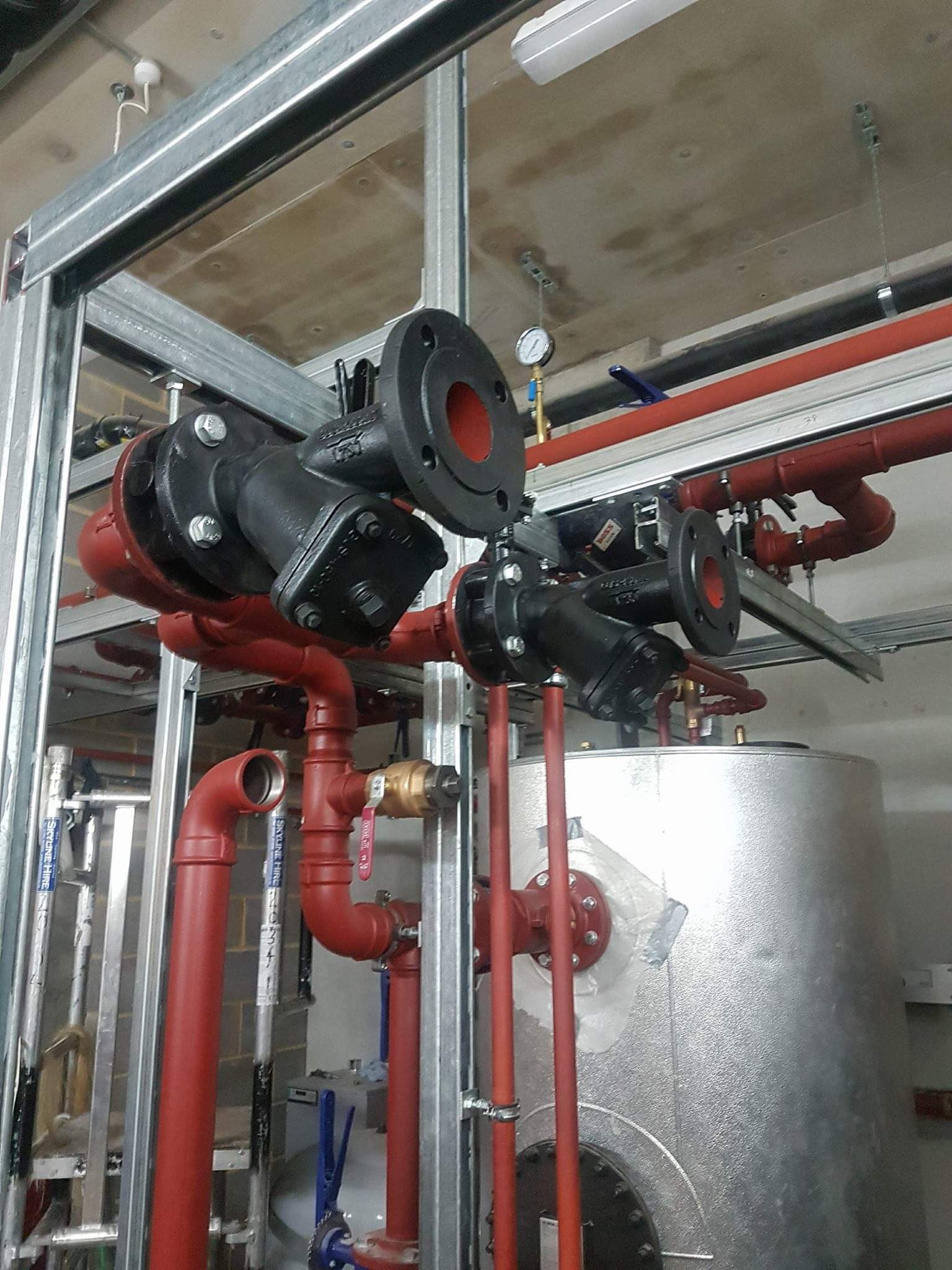 Czerwone rury instalacji grzewczej z czarnymi zaworami i srebrnym zbiornikiem, zamontowane w budynku w trakcie budowy, widoczne metalowe profile konstrukcyjne.
