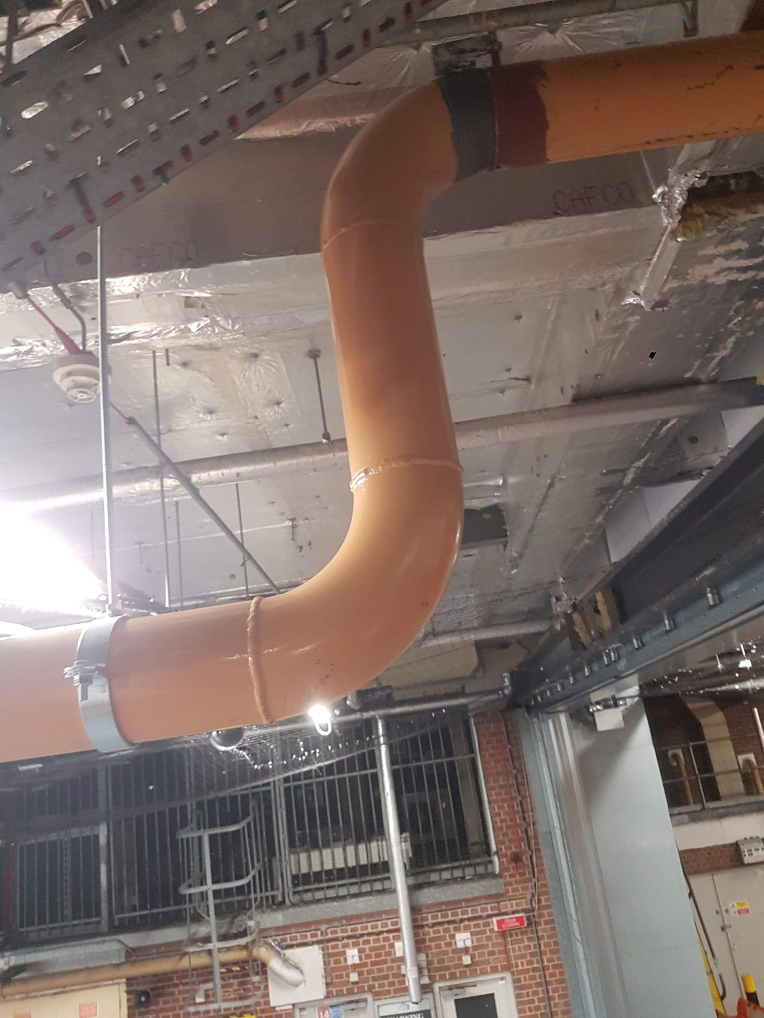 Pomarańczowa rura kanalizacyjna z obejmą, zamontowana pod sufitem w budynku przemysłowym. Widoczne elementy instalacji, sufit z izolacją i fragment ściany z oknami.