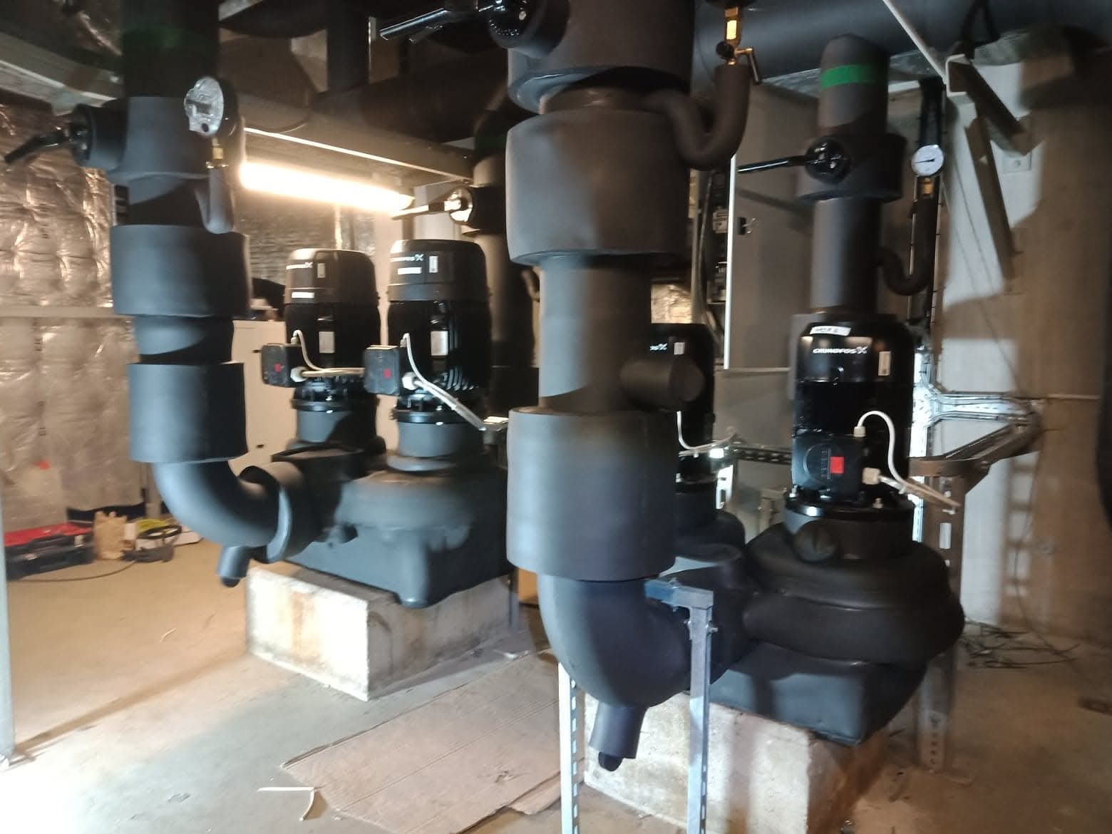 Czarne pompy obiegowe Grundfos w kotłowni, zaizolowane rury, manometry, instalacja na betonowych podstawach, oświetlenie jarzeniowe.