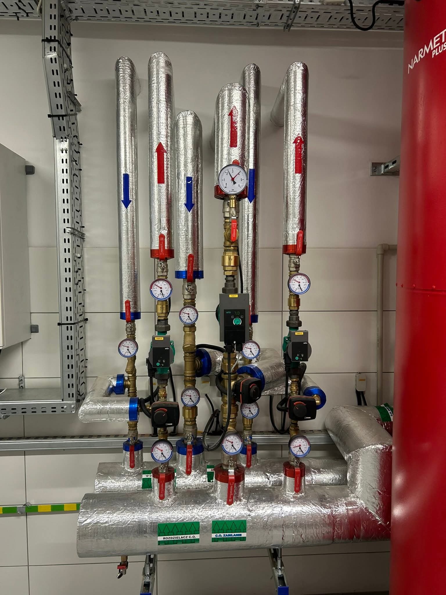 Izolowane rury instalacji grzewczej z manometrami i zaworami, widoczne strzałki kierunku przepływu, na tle białych płytek i czerwonego zbiornika.