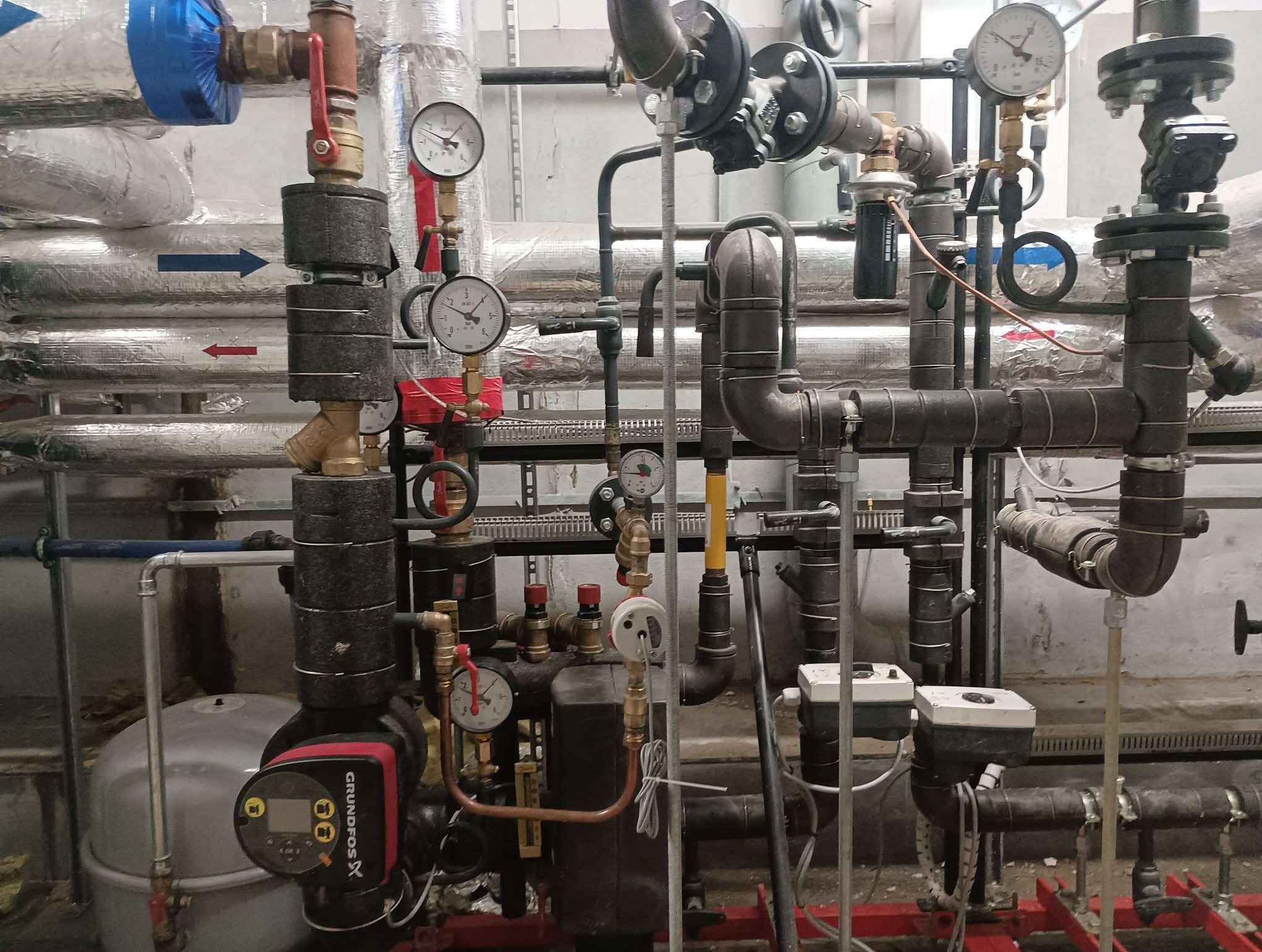 Złożona instalacja grzewcza z rurami, zaworami, manometrami i pompą Grundfos. Rury izolowane termicznie, oznaczone strzałkami kierunku przepływu. Widoczne elementy mocujące i podtrzymujące.