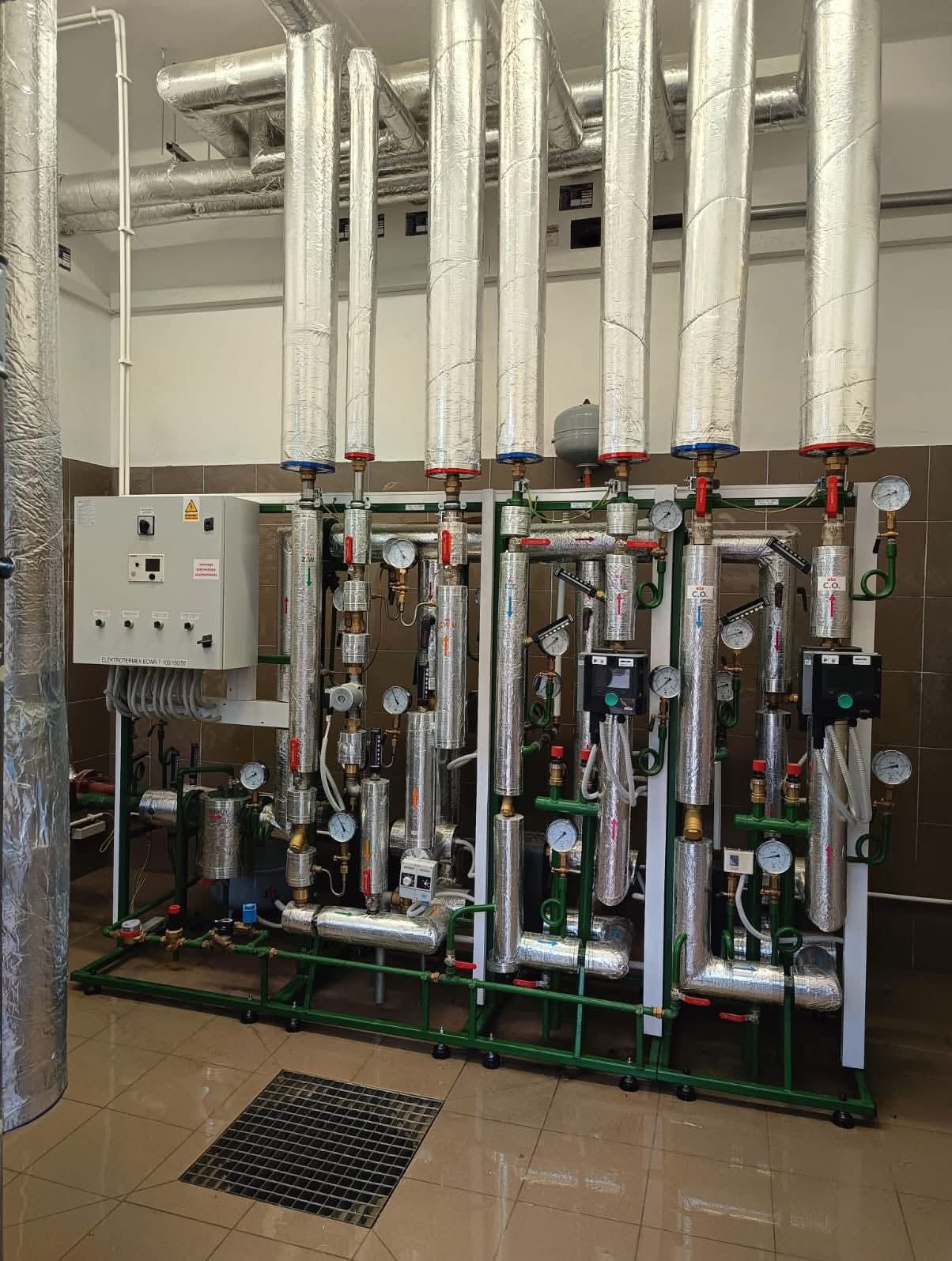 Złożona instalacja grzewcza z rurami w izolacji, manometrami i zaworami, umieszczona w pomieszczeniu z brązowymi płytkami i kratką ściekową w podłodze. Widoczna szafka sterownicza.