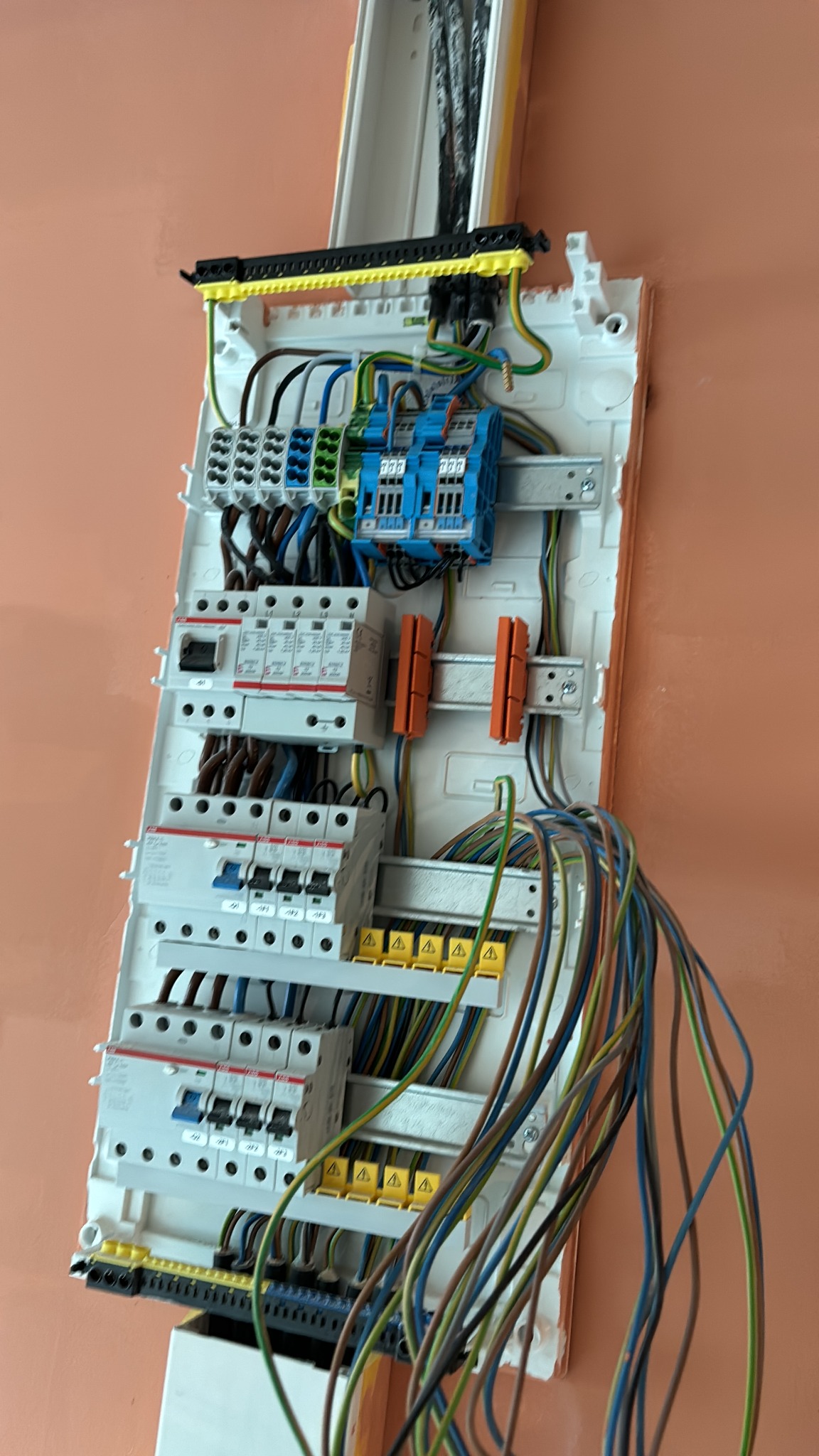 Rozdzielnica elektryczna z zabezpieczeniami przepięciowymi i różnicowoprądowymi, widok z góry, kable w różnych kolorach, pomarańczowa ściana w tle.