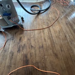 GEKA Podłogi Emil Wierzbowski - Cyklinowanie parkietu dębowego maszyną z odkurzaczem. Widoczne przewody i wąż ssący. Podłoga po obróbce, z wyraźnym rysunkiem drewna i matowym wykończeniem.