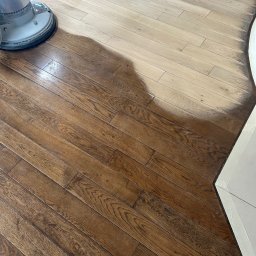 GEKA Podłogi Emil Wierzbowski - Cyklinowanie parkietu w trakcie realizacji: maszyna do cyklinowania obok fragmentu odnowionej, ciemnej podłogi z parkietu, kontrastującej z jasnym, nieobrobionym obszarem.