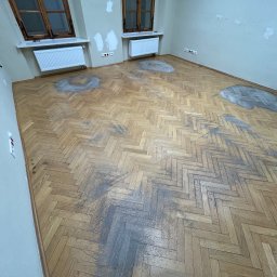 GEKA Podłogi Emil Wierzbowski - Parkiet ułożony w jodełkę przed cyklinowaniem. Widoczne plamy i zabrudzenia na drewnianej podłodze. Dwa okna z drewnianą ramą i grzejniki pod nimi.