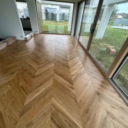 GEKA Podłogi Emil Wierzbowski - Jasny parkiet ułożony we wzór jodełki francuskiej w przestronnym wnętrzu z dużymi oknami. Na podłodze leżą jeszcze nieużyte deski.