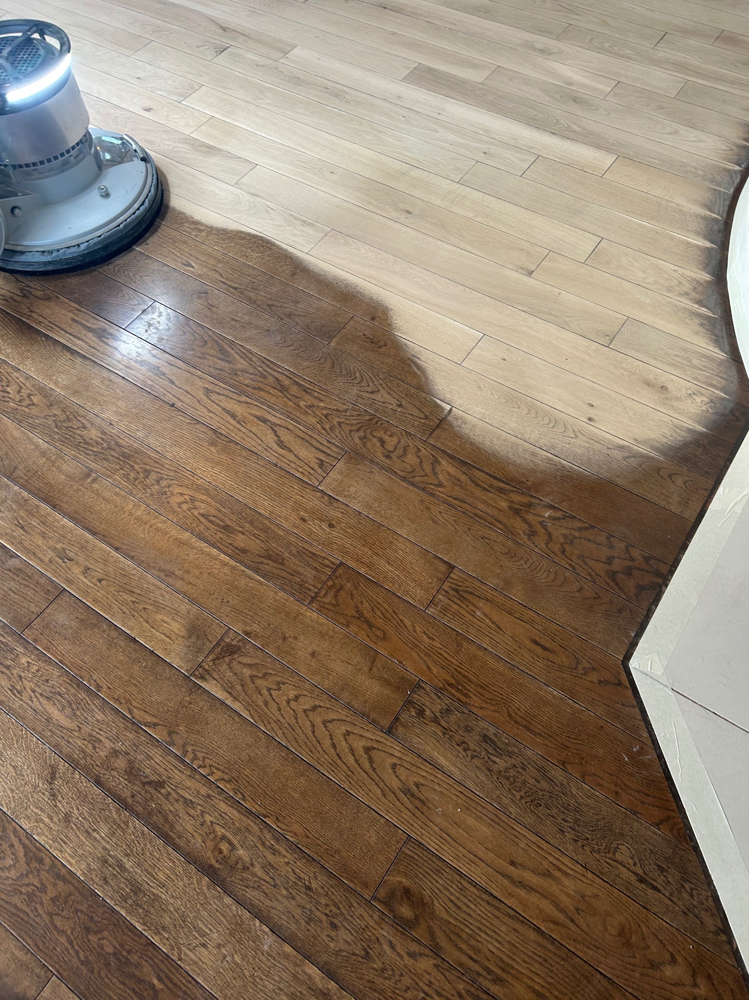 Cyklinowanie parkietu w trakcie realizacji: maszyna do cyklinowania obok fragmentu odnowionej, ciemnej podłogi z parkietu, kontrastującej z jasnym, nieobrobionym obszarem.