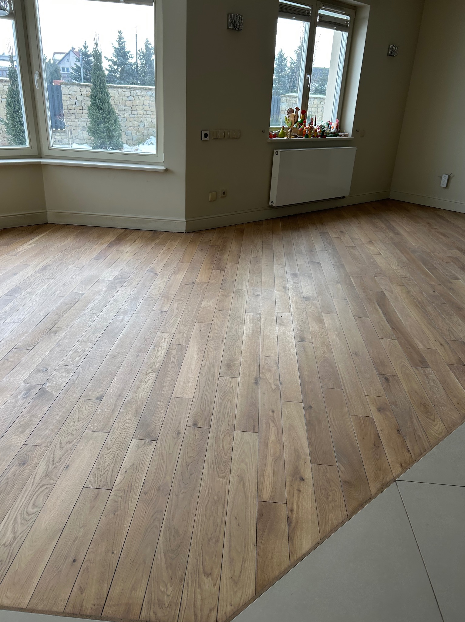 Jasne, drewniane panele ułożone pod kątem w przestronnym pokoju z oknami. Widoczne dekoracje na parapecie i fragment podłogi wyłożonej płytkami.