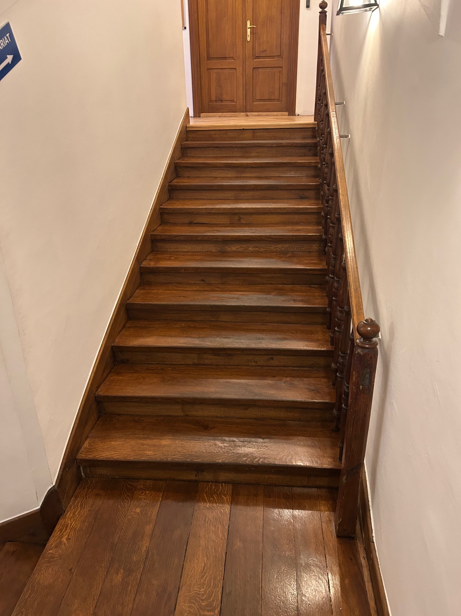 Drewniane schody z poręczą w klasycznym stylu, widok z dołu. Parkiet na podeście, białe ściany, drzwi na górze. Ciepłe oświetlenie podkreśla rysunek drewna.