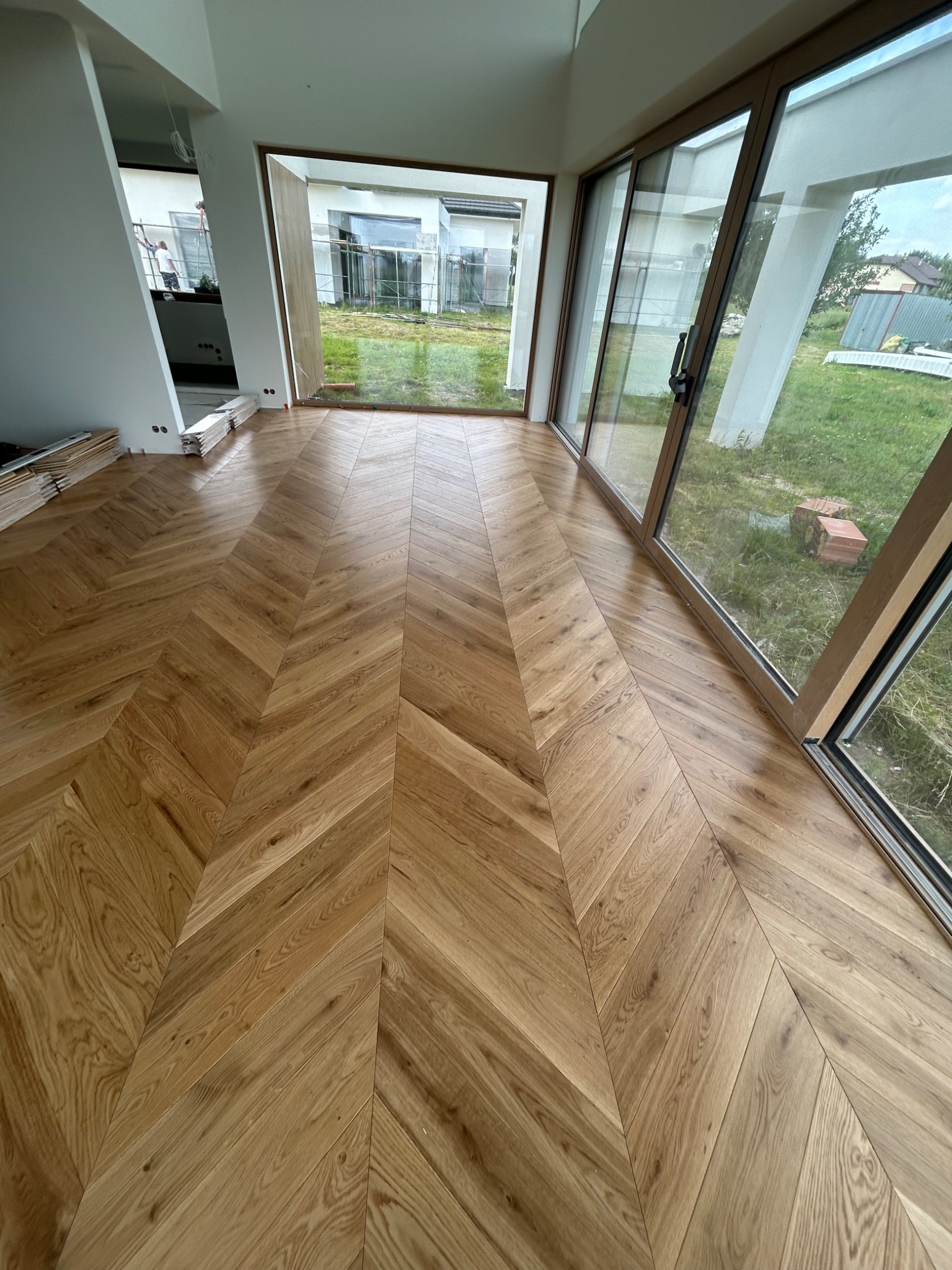 Jasny parkiet ułożony we wzór jodełki francuskiej w przestronnym wnętrzu z dużymi oknami. Na podłodze leżą jeszcze nieużyte deski.