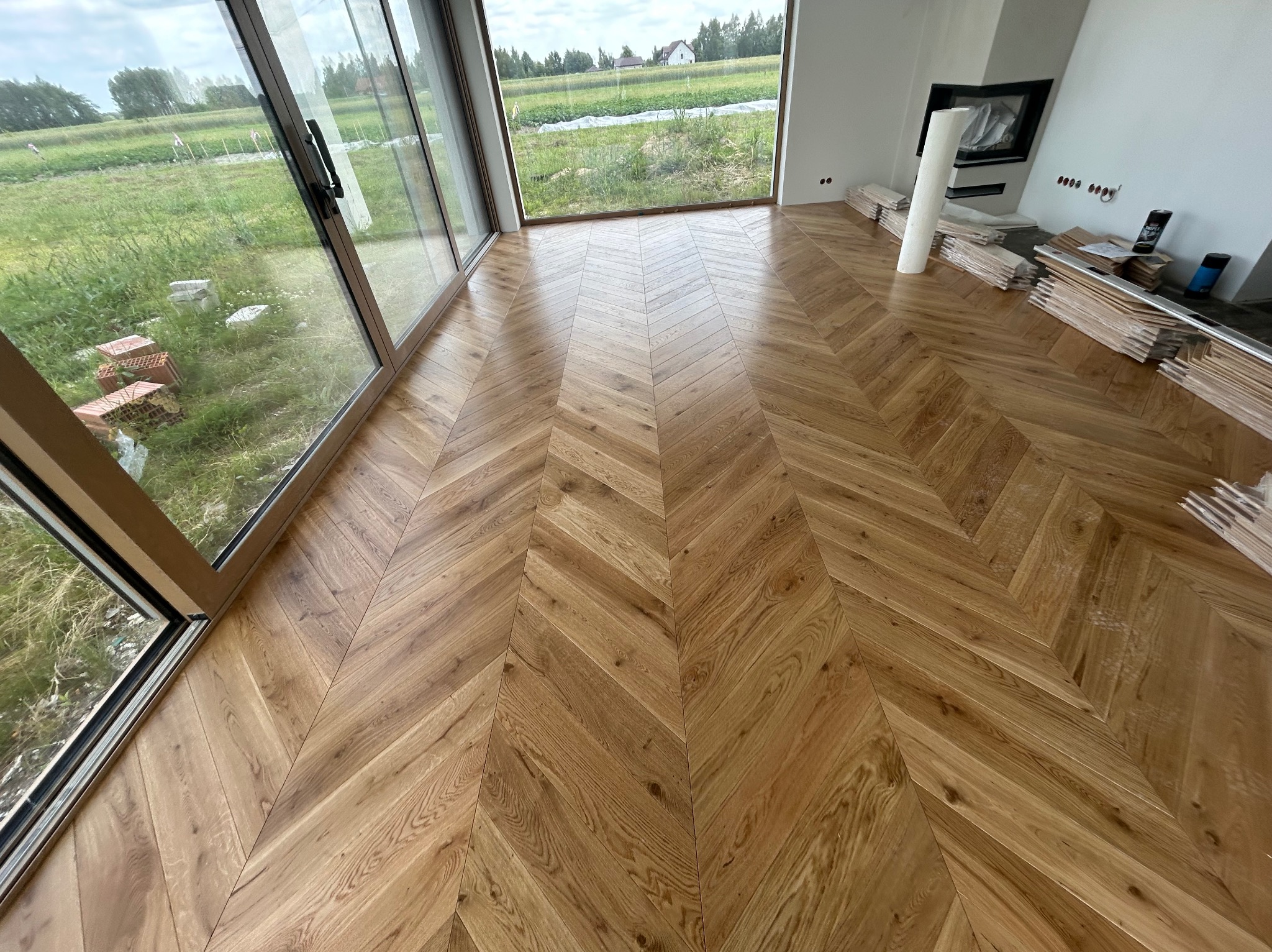 Ułożony parkiet w jodełkę francuską, widok na duże okno z zielonym ogrodem. Część desek i akcesoriów do montażu leży obok kominka. Jasne wnętrze, naturalne światło.