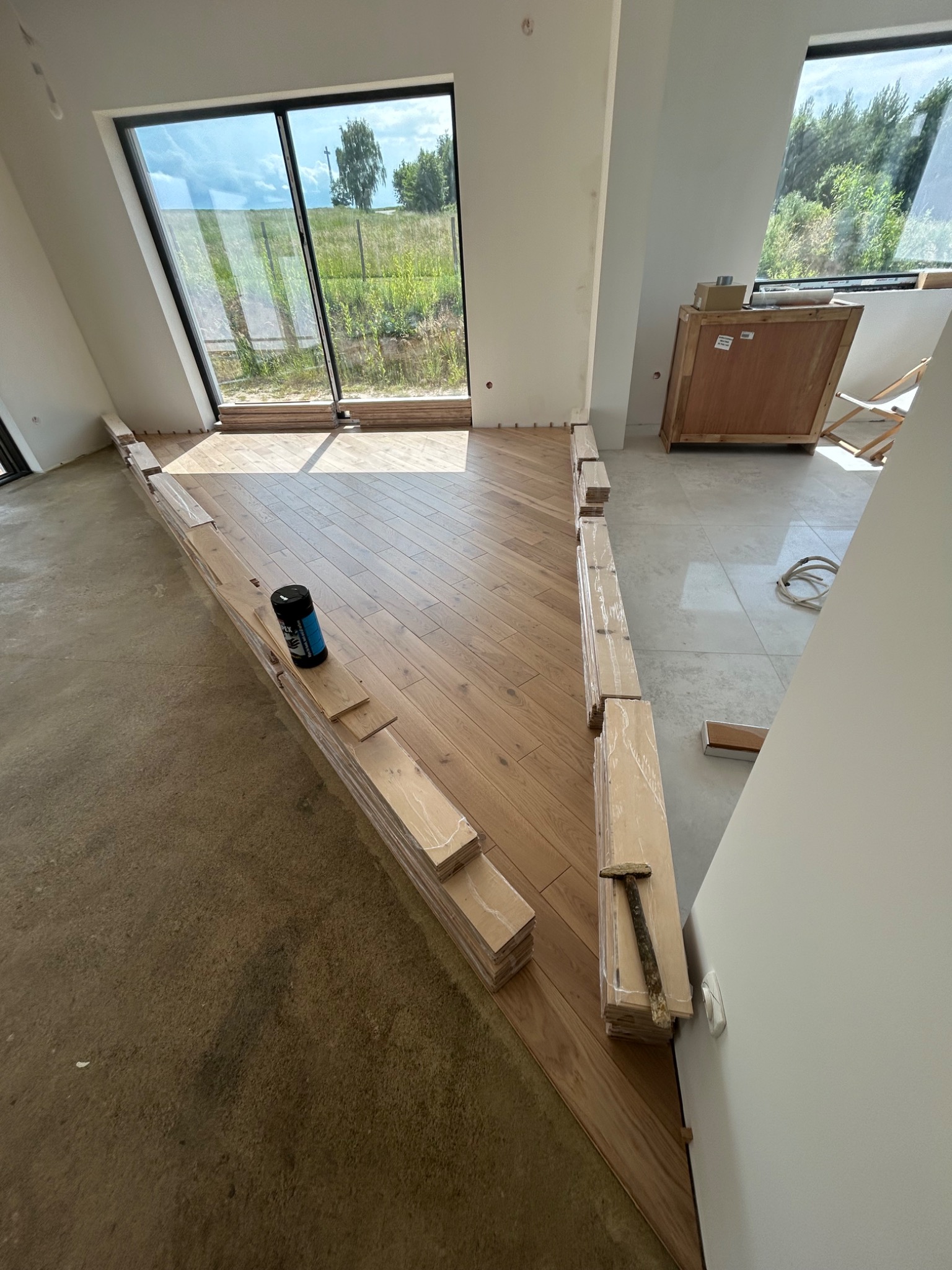 Układanie parkietu w jodełkę francuską w jasnym wnętrzu z dużym oknem. Częściowo ułożona podłoga, paczki parkietu i narzędzia. Widok na zewnątrz.