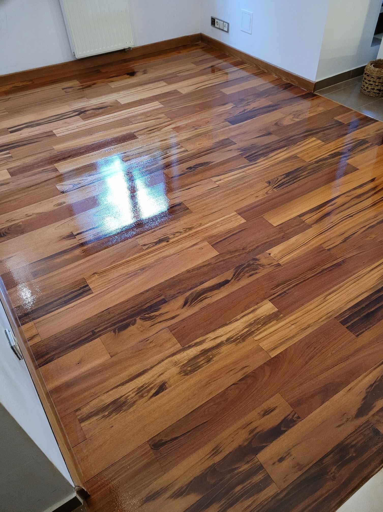 Połyskująca, świeżo ułożona podłoga z parkietu o egzotycznym wzorze drewna, z widocznymi refleksami światła i listwami przypodłogowymi w kolorze drewna.