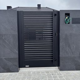 NewDecor Krzysztof Dobrowolski - Nowoczesna, czarna brama wjazdowa z poziomymi szczeblami, zamontowana w szarym, kamiennym ogrodzeniu. Widoczne przewody instalacji elektrycznej.