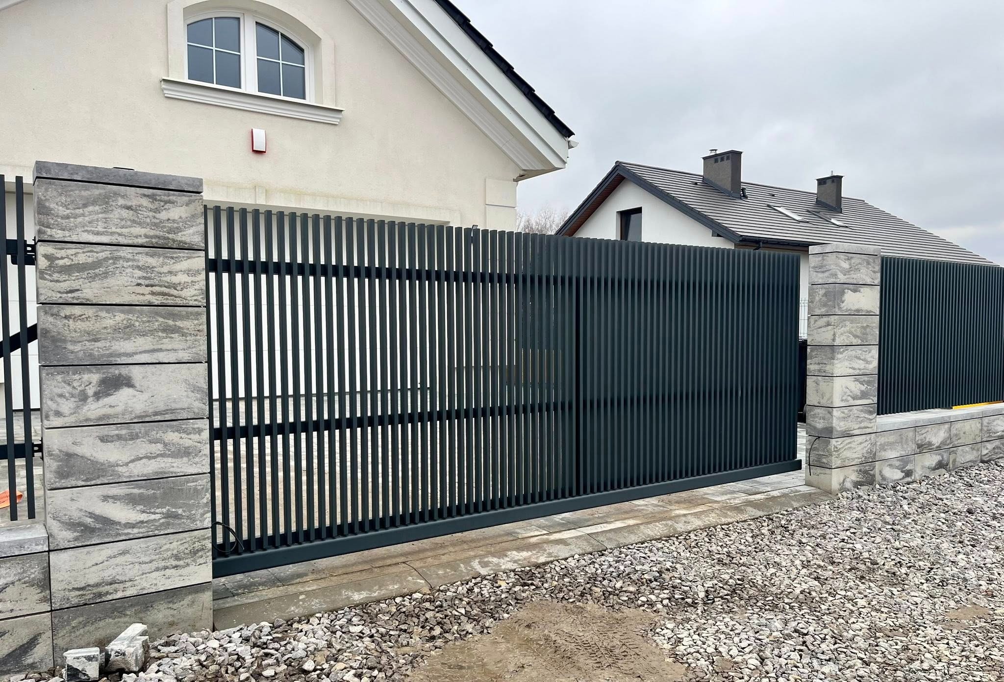 Nowoczesna brama przesuwna z pionowych profili, osadzona na słupkach z szarej kostki w Zalesiu Górnym. Minimalistyczny design, jasna elewacja domu w tle.