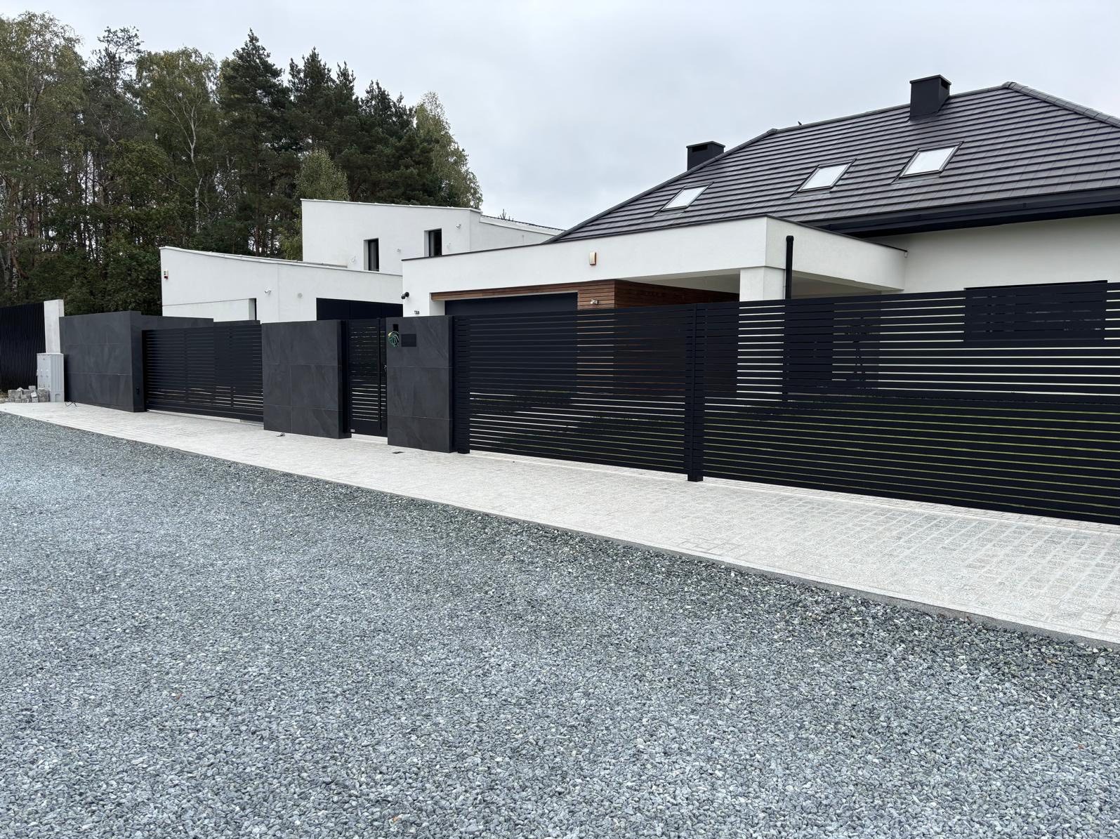 Nowoczesne, poziome, czarne ogrodzenie z bramą wkomponowane w architekturę domu. Szara kostka brukowa i żwir na podjeździe. Minimalistyczny design.