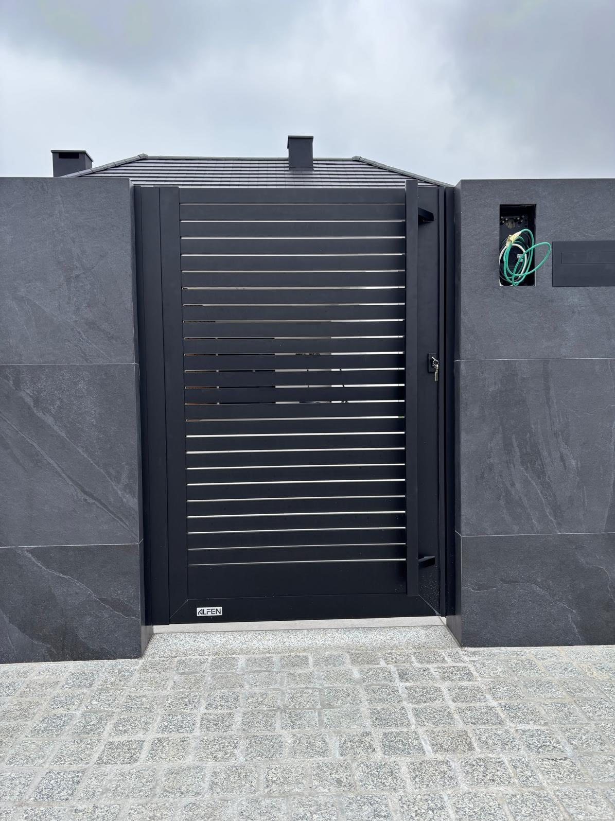 Nowoczesna, czarna brama wjazdowa z poziomymi szczeblami, zamontowana w szarym, kamiennym ogrodzeniu. Widoczne przewody instalacji elektrycznej.