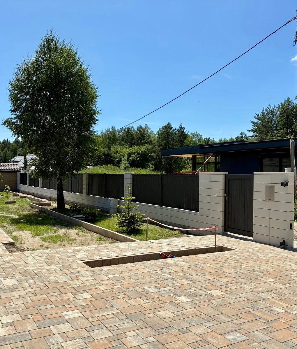 Nowoczesne ogrodzenie panelowe z bramą w kolorze antracytowym, osadzone na betonowej podmurówce. Widok na posesję z kostką brukową i zielenią w tle.