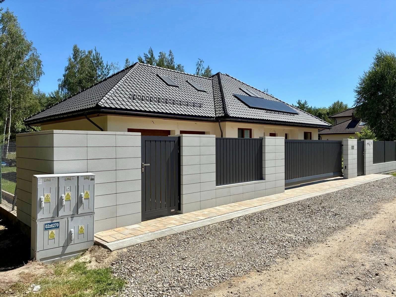 Nowoczesna posesja z przesuwną bramą i furtką w grafitowej kolorystyce, osadzona w betonowych słupkach. Widoczne panele słoneczne na dachu domu.